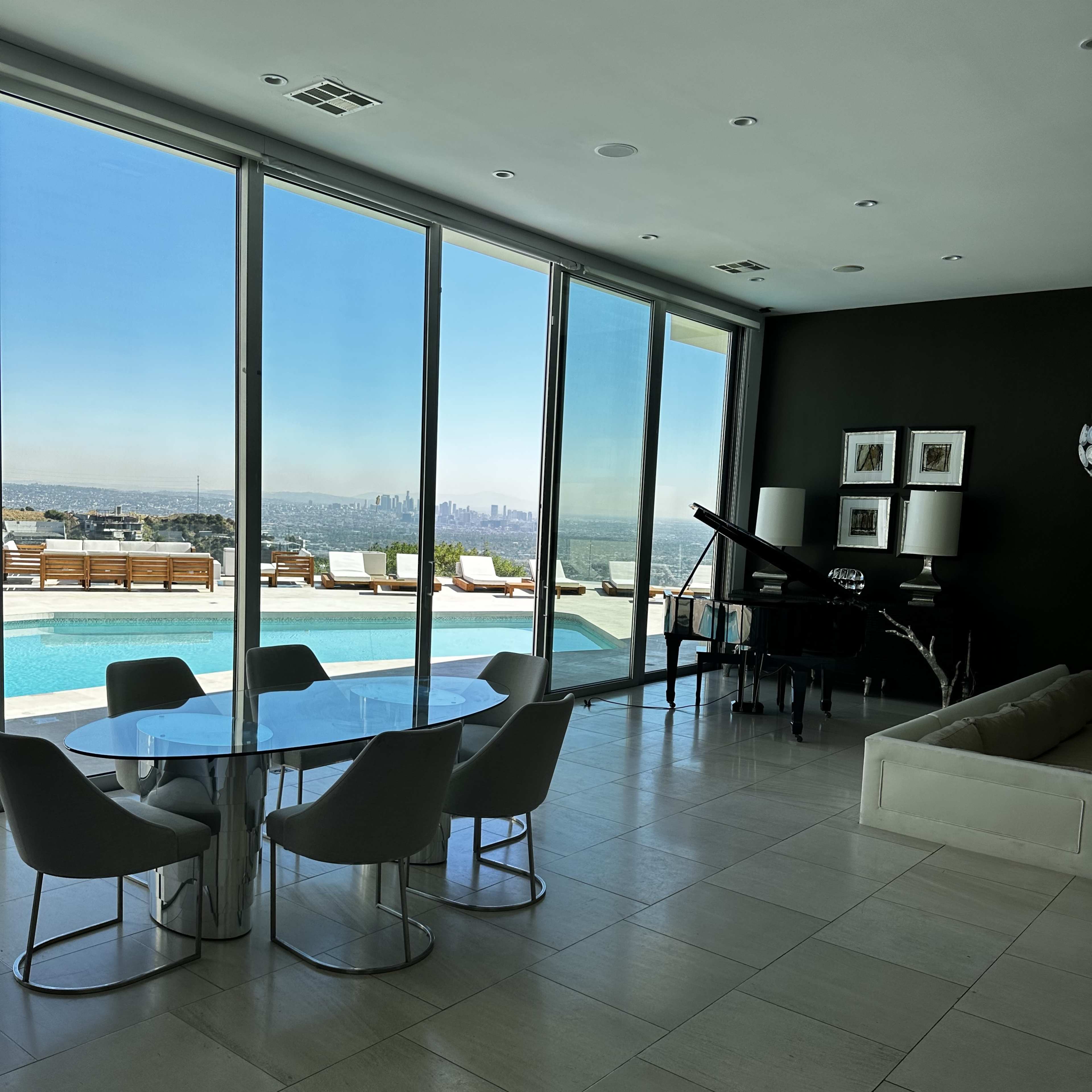 A modern interior space featuring a round dining table with six chairs, a grand piano, and large windows overlooking a pool and city skyline.