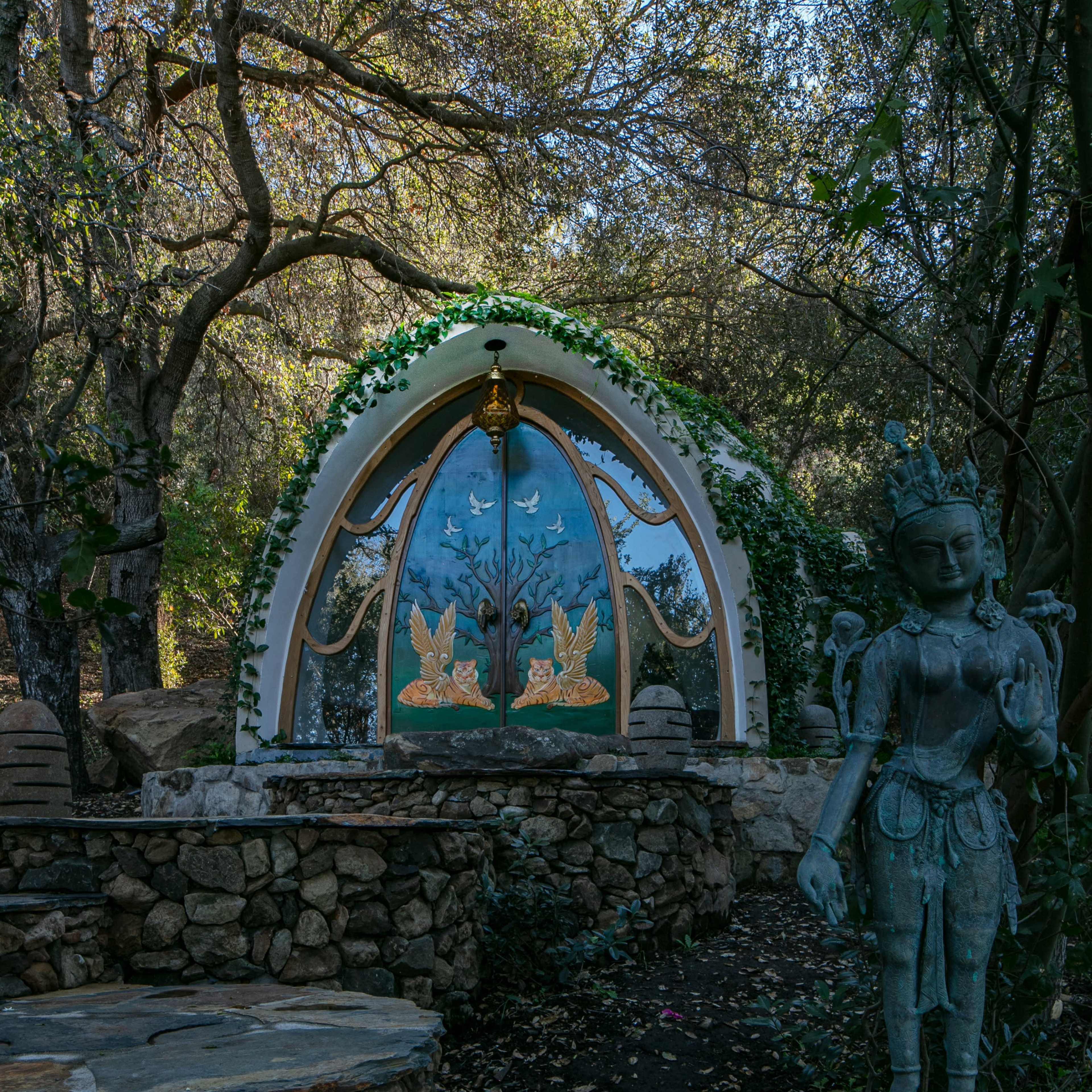 A stone path leads to a decorated entrance featuring a colorful arch with murals and a statue beside it in a wooded setting.