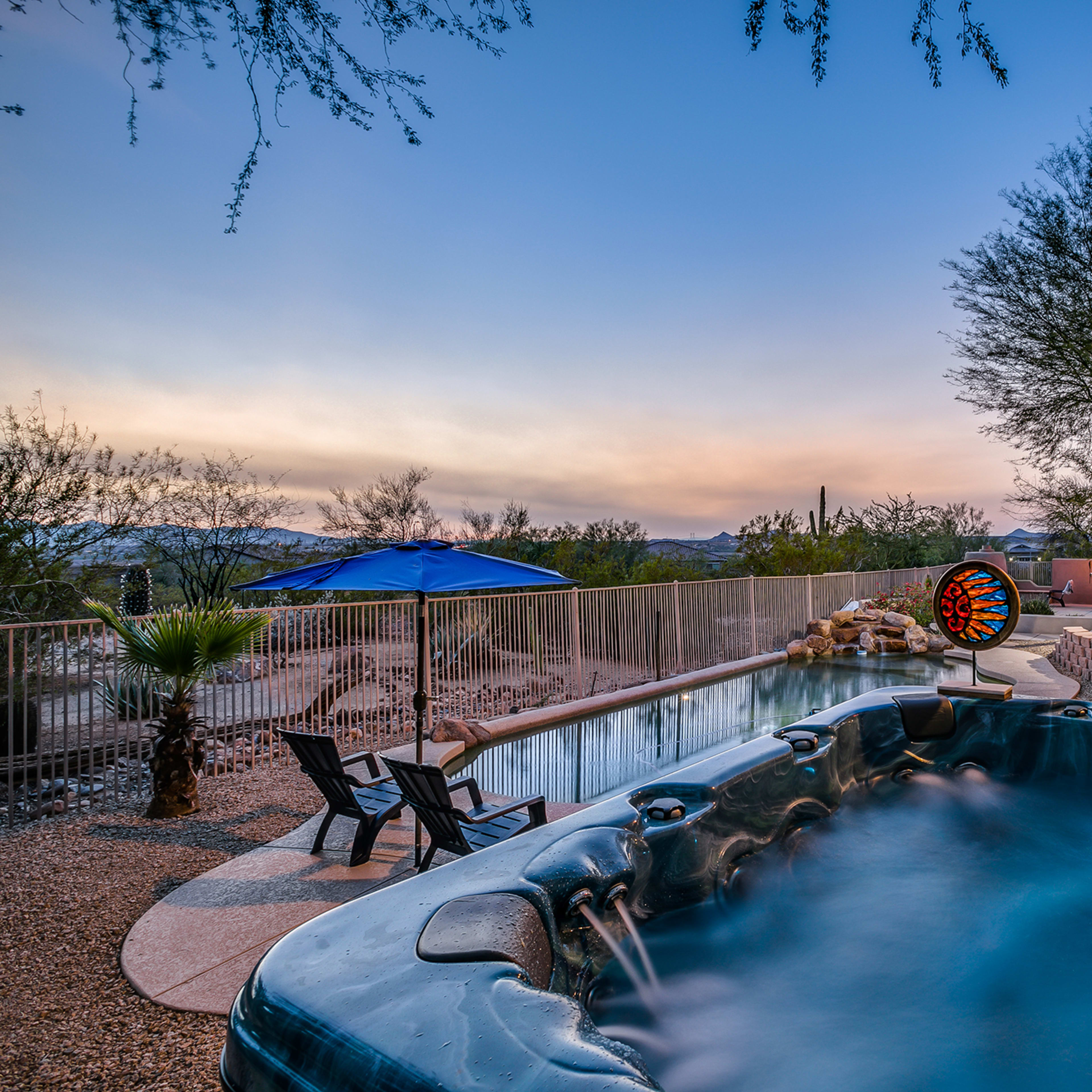 A hot tub beside a swimming pool, surrounded by desert landscaping and outdoor seating, under a twilight sky.