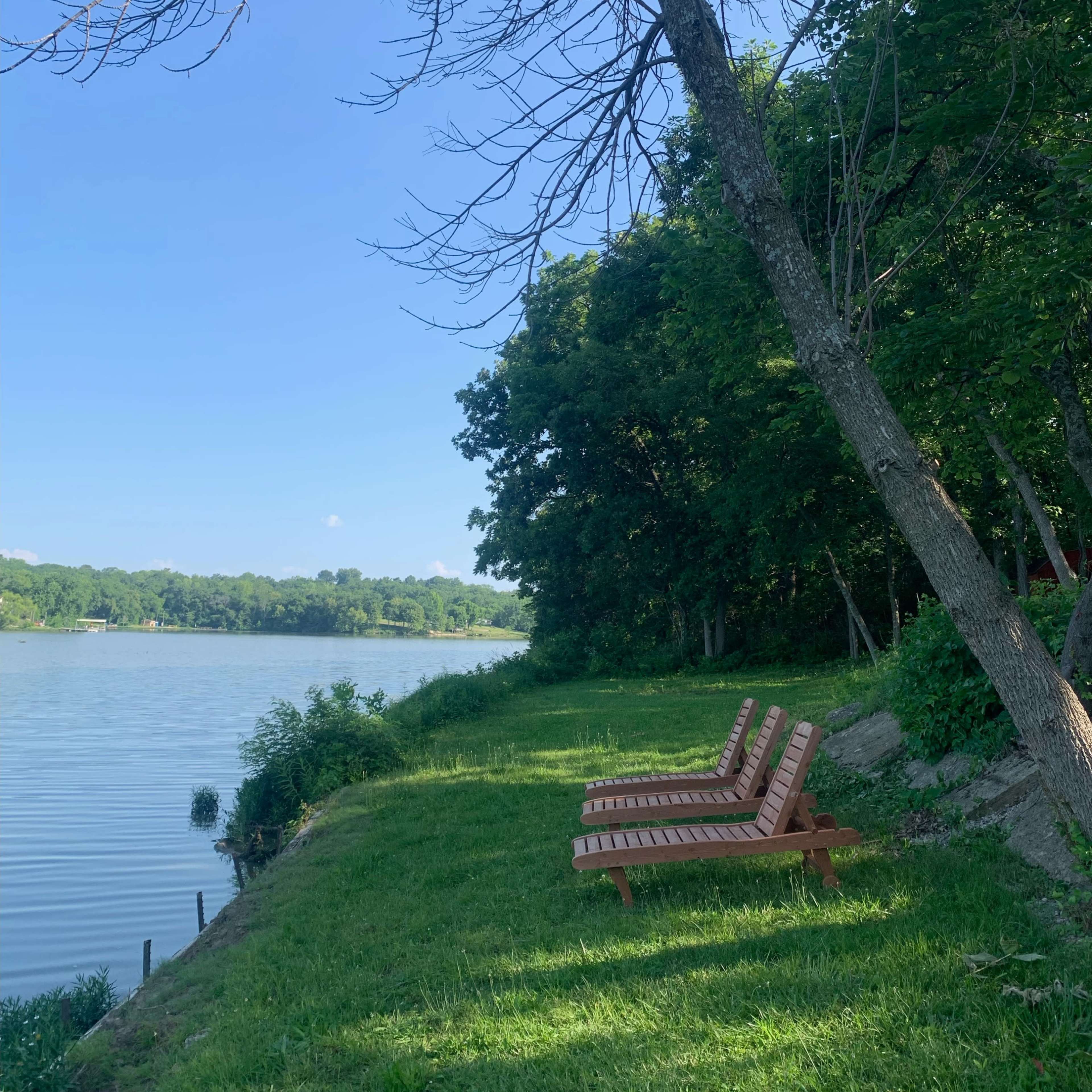 Two wooden lounge chairs positioned on a grassy shore beside a calm river, with trees and a clear blue sky in the background.