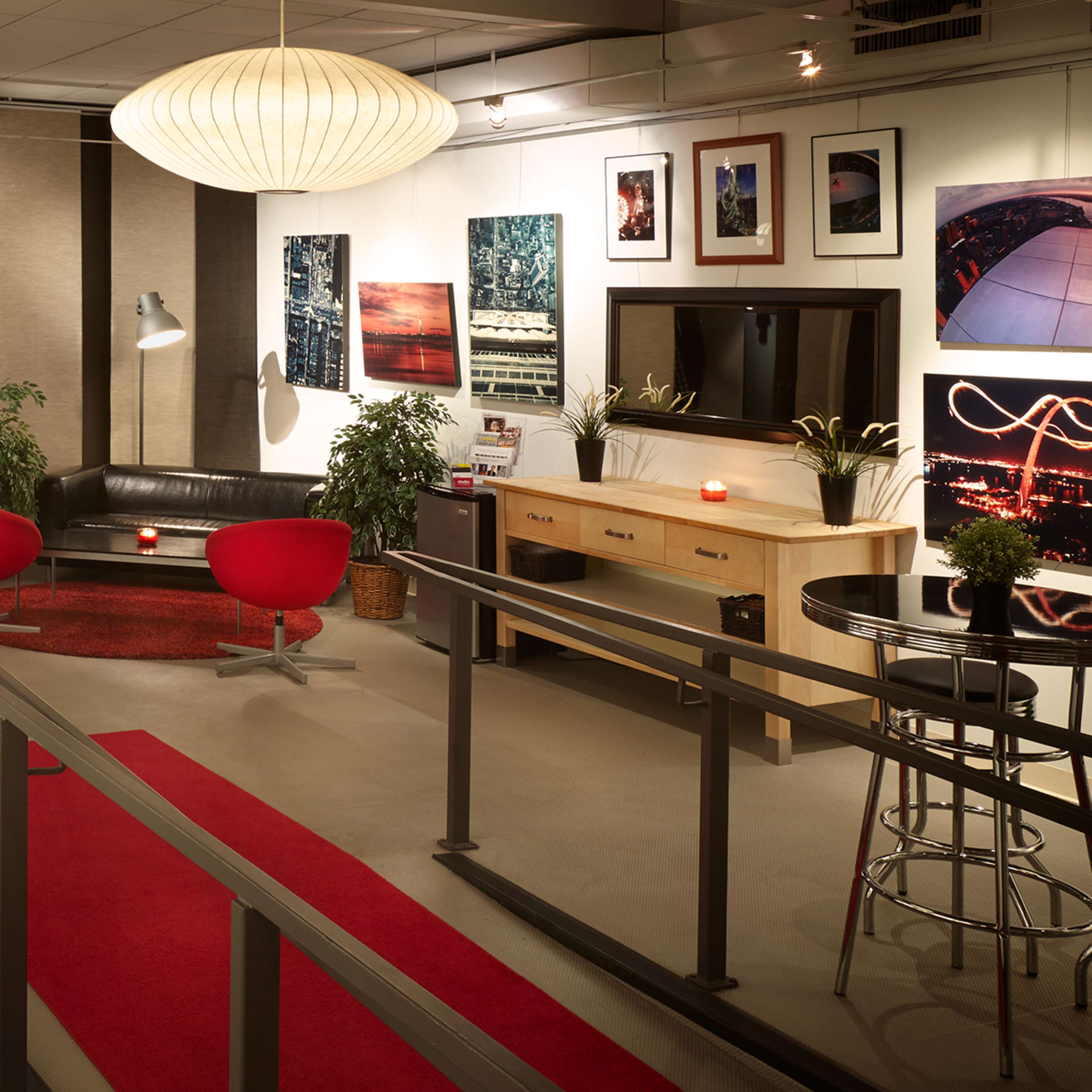 A modern lounge area with a black sofa, red accent chairs, a coffee table, and framed photos on the walls, illuminated by pendant lighting.