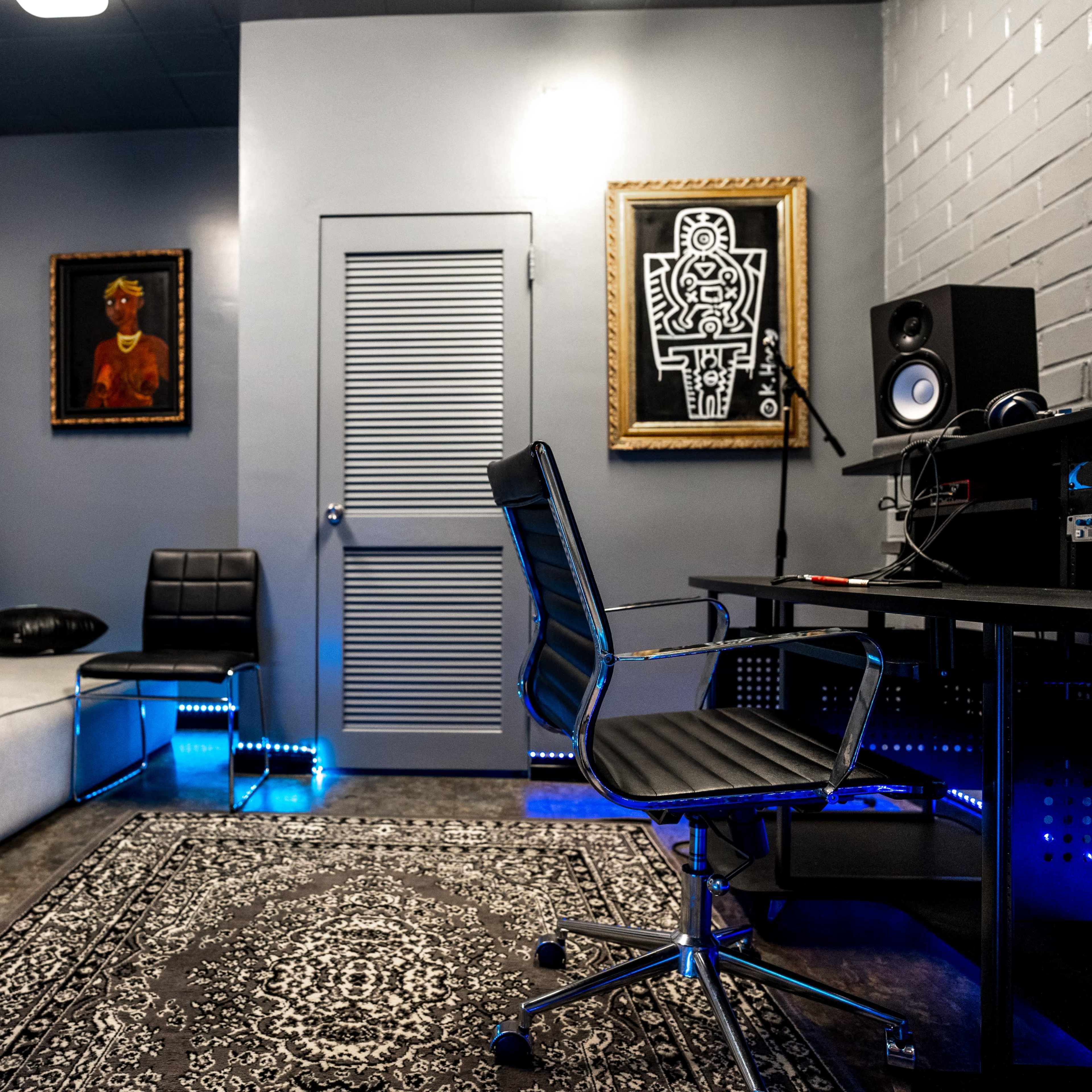 A modern, minimalist room featuring a gray sofa, a black chair, a desk with a computer setup, and artwork on the walls.