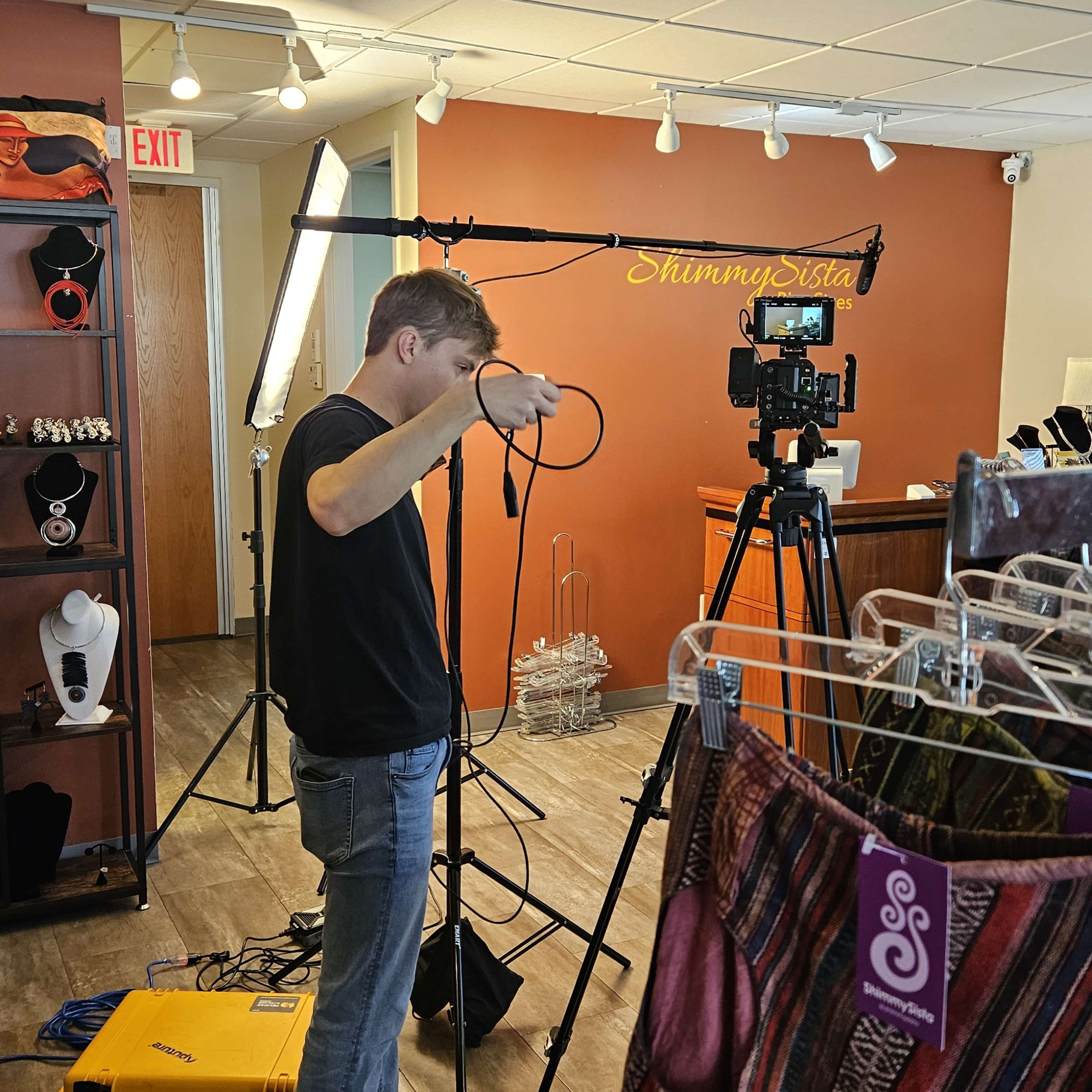 A person is setting up camera equipment and organizing cables in a retail space with a backdrop sign that reads "Shimmering Style."
