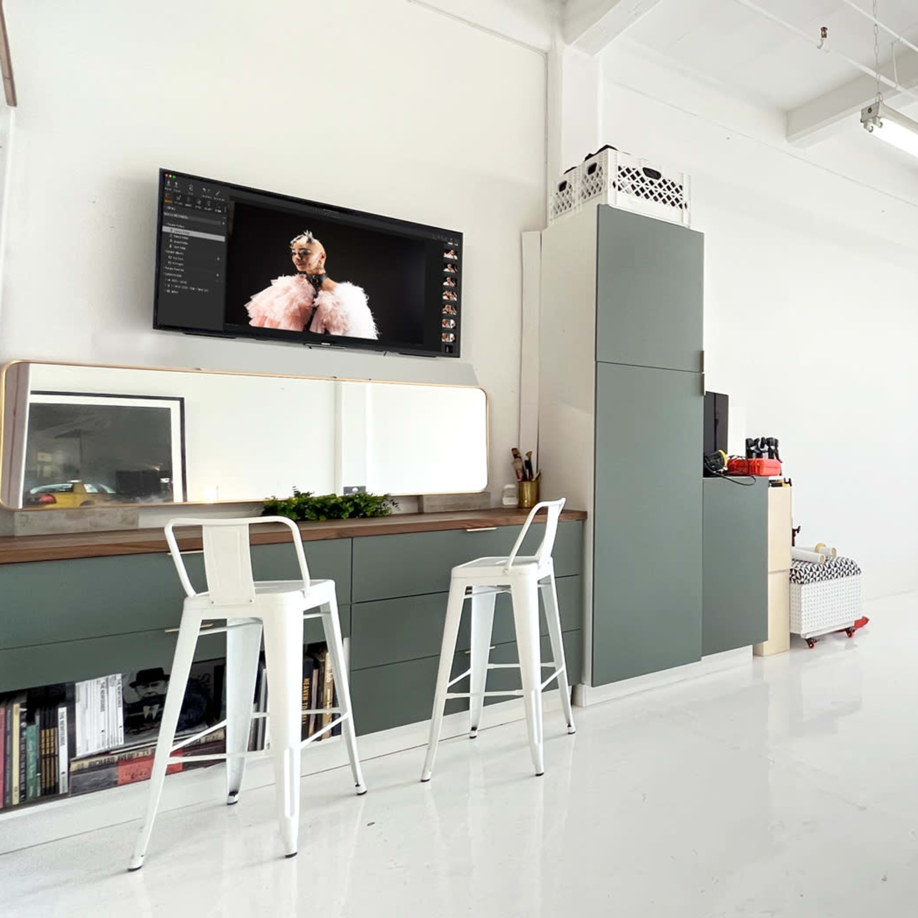 A bright, minimalist studio with a large window, two white stools at a green vanity, and a television displaying an image on the wall.