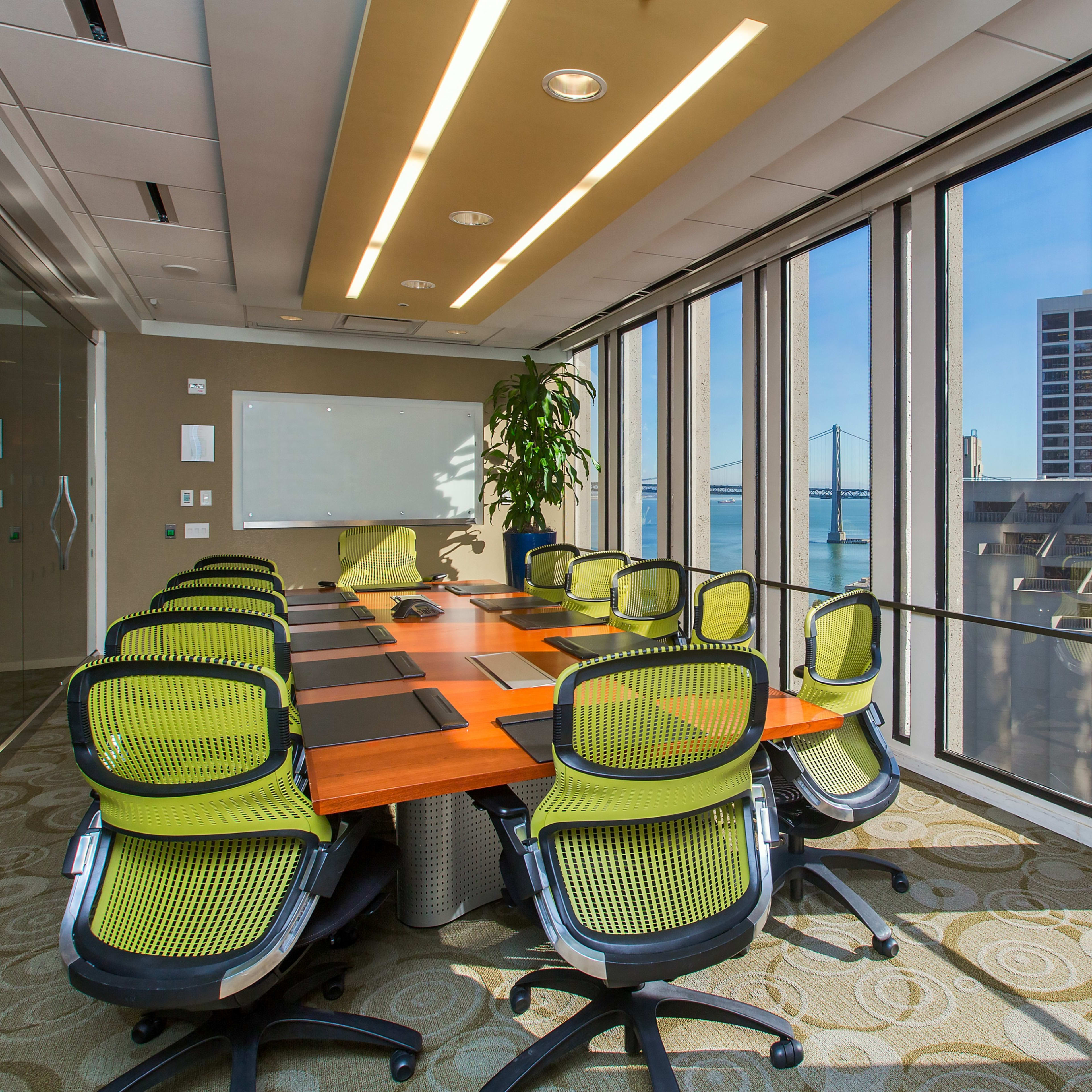 A modern conference room features a large wooden table surrounded by green office chairs and floor-to-ceiling windows overlooking a cityscape and water.