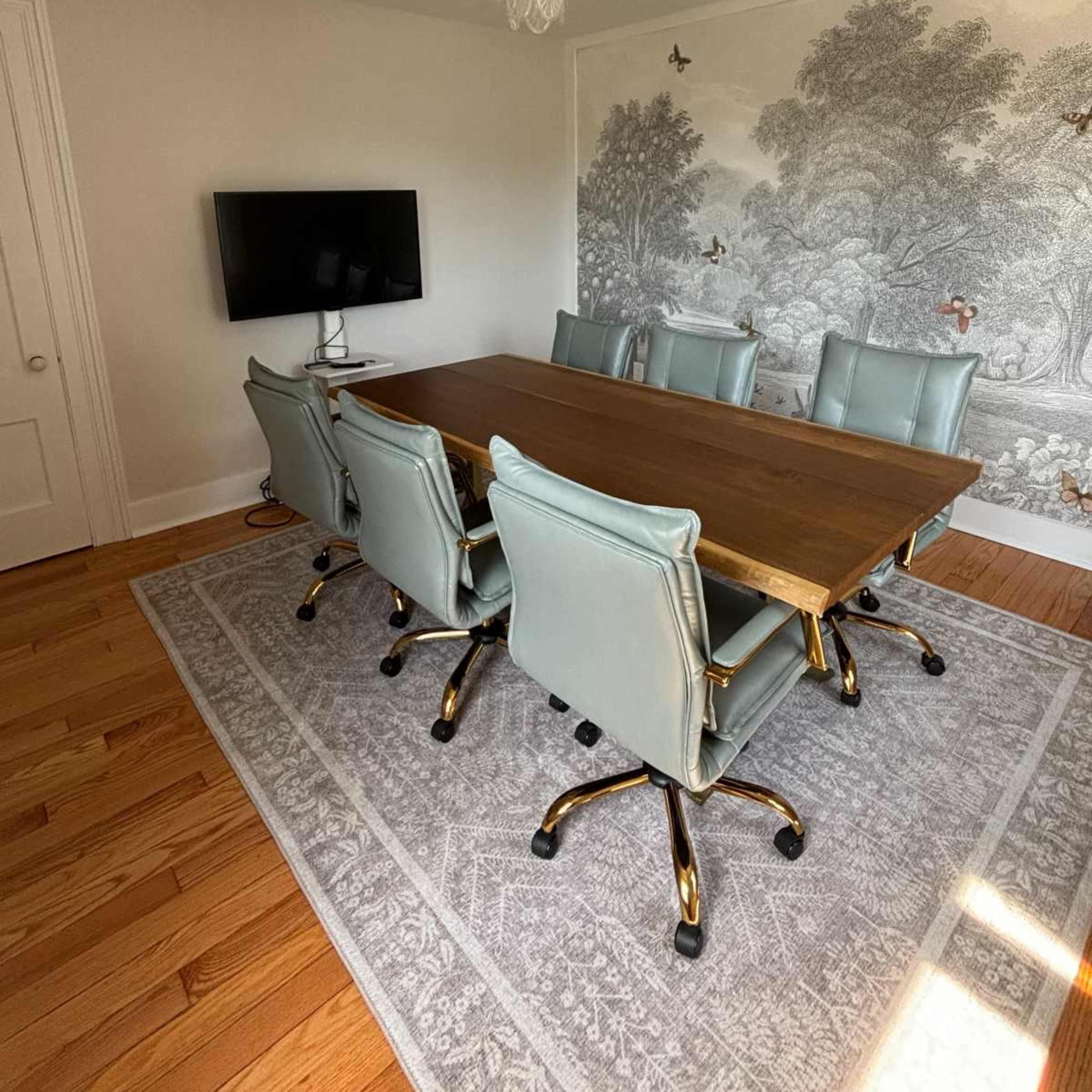 A modern conference room with a wooden table surrounded by six light blue office chairs, a wall-mounted TV, and a decorative mural depicting a landscape.