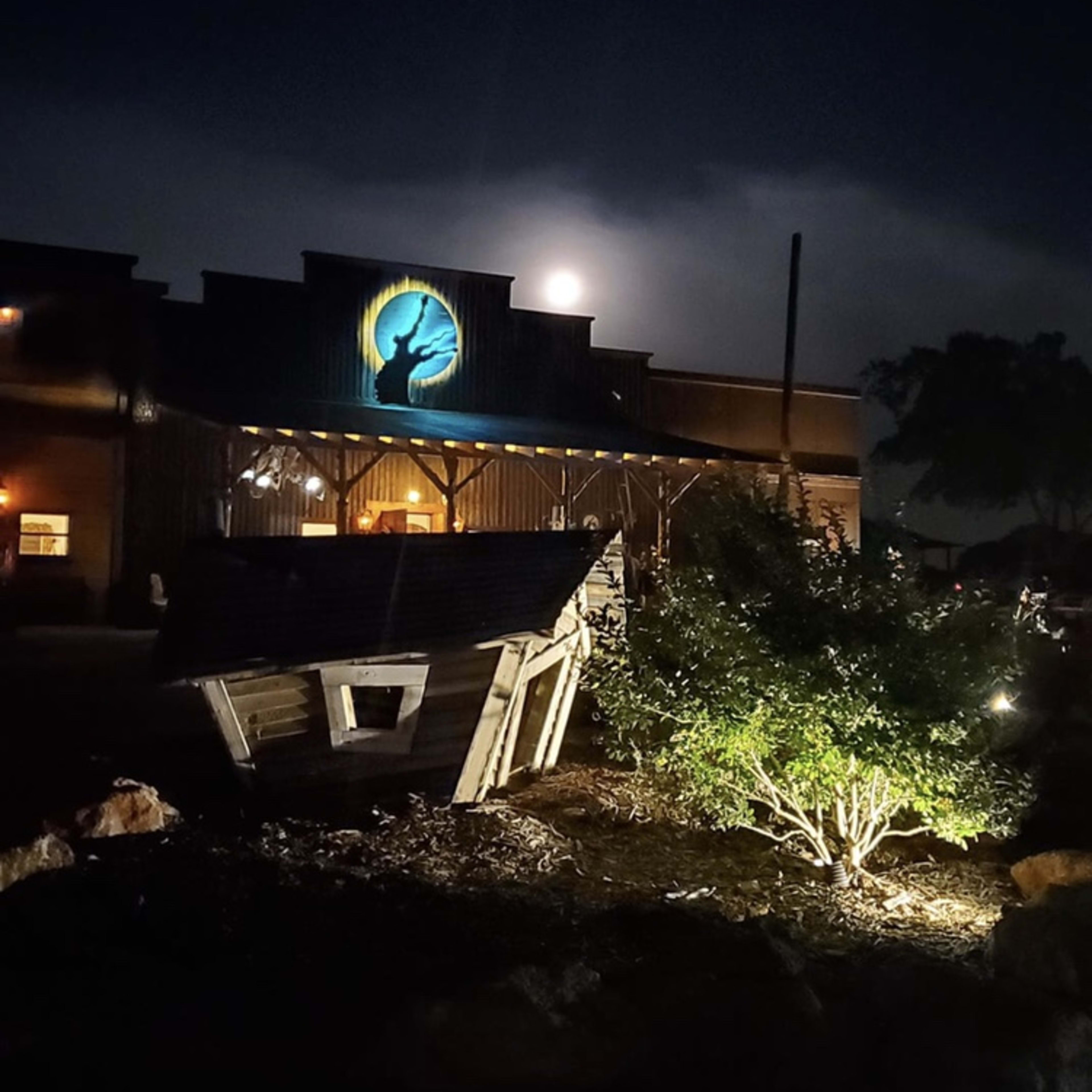 A full moon illuminates the facade of a rustic building, while a wooden sign and shrubbery are lit by ground lights in the foreground.