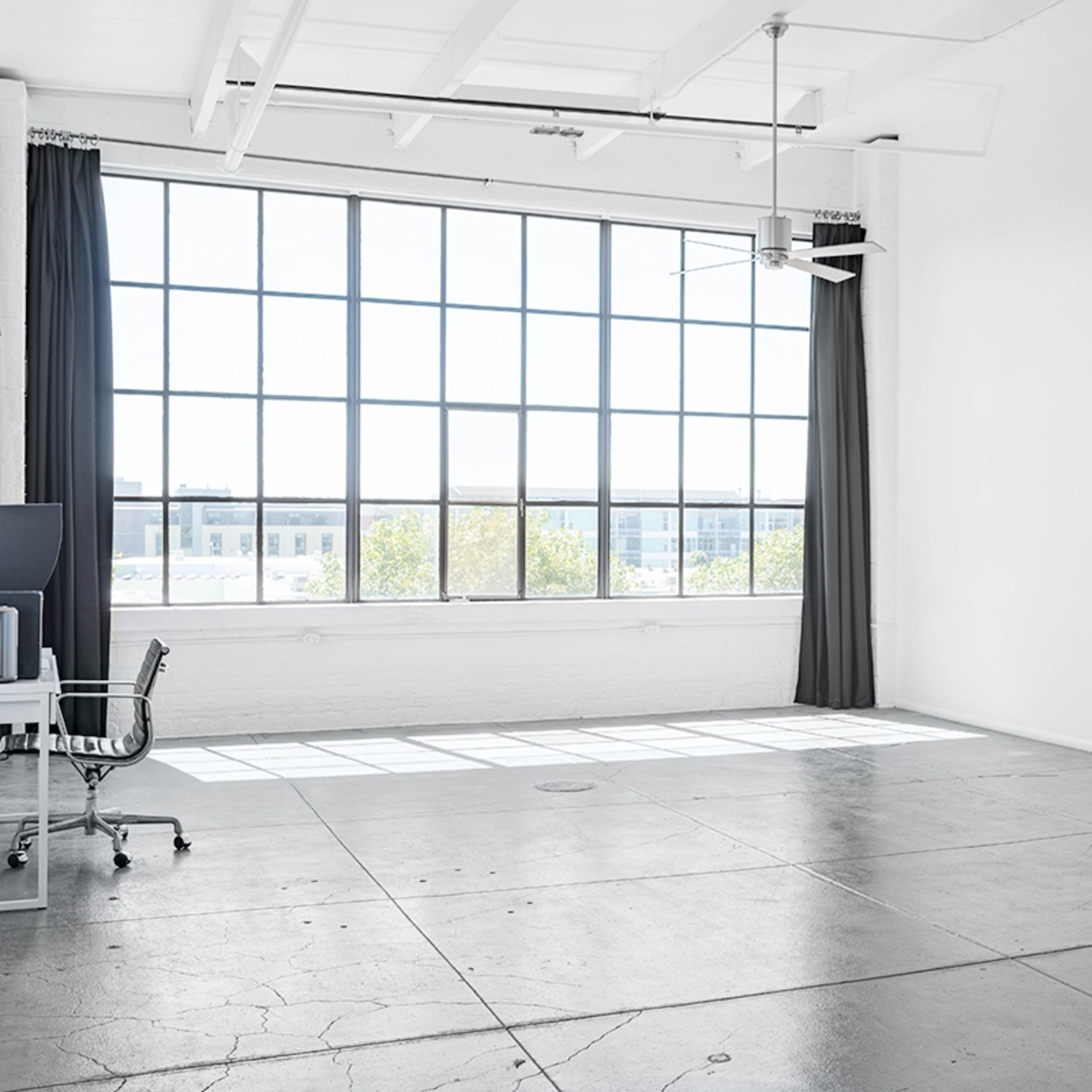 A bright, spacious room features a large window with a view, a ceiling fan, and minimal furniture including a chair and a stand.