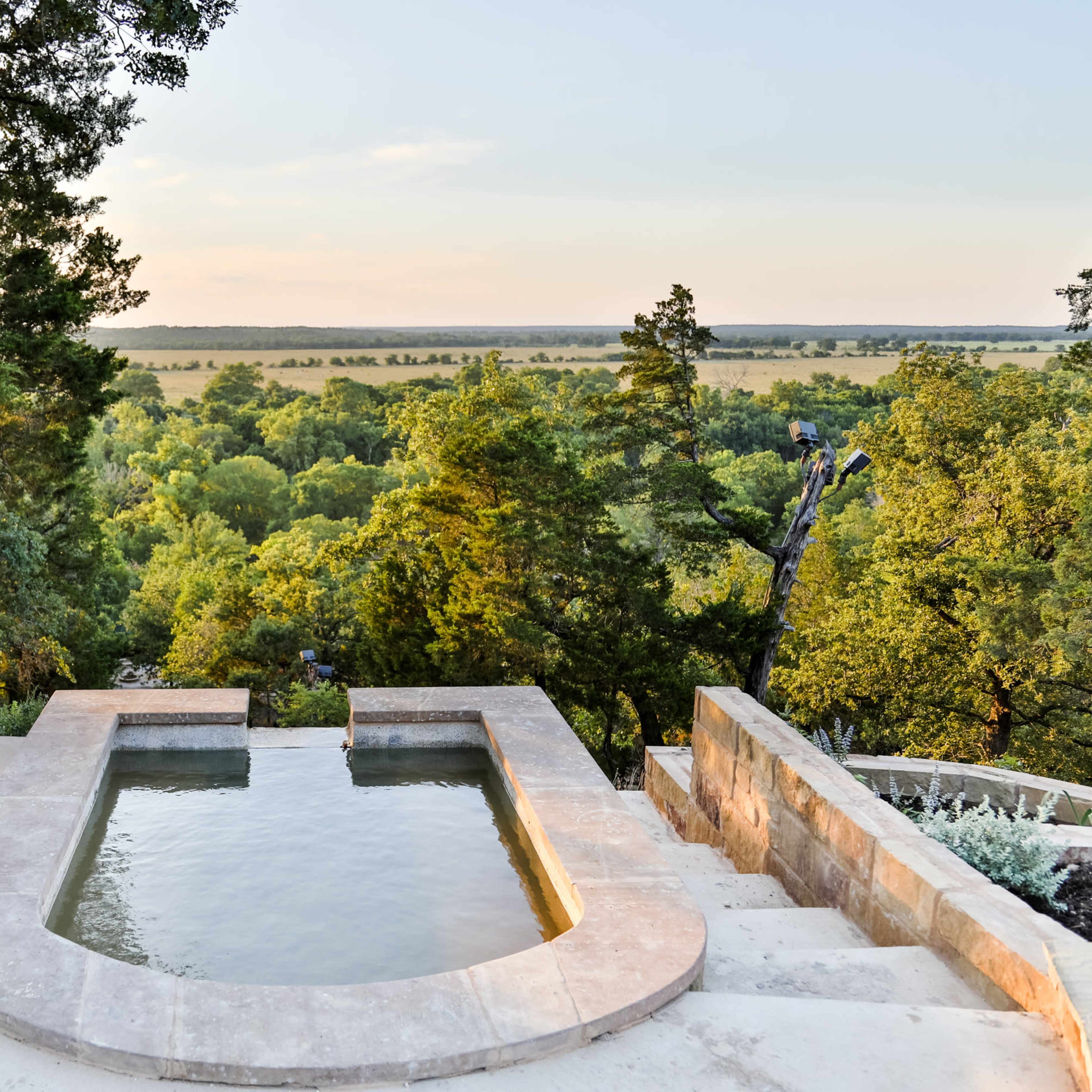 A rectangular, stone-lined pool is situated on a hillside, overlooking a lush green landscape with trees and rolling fields.