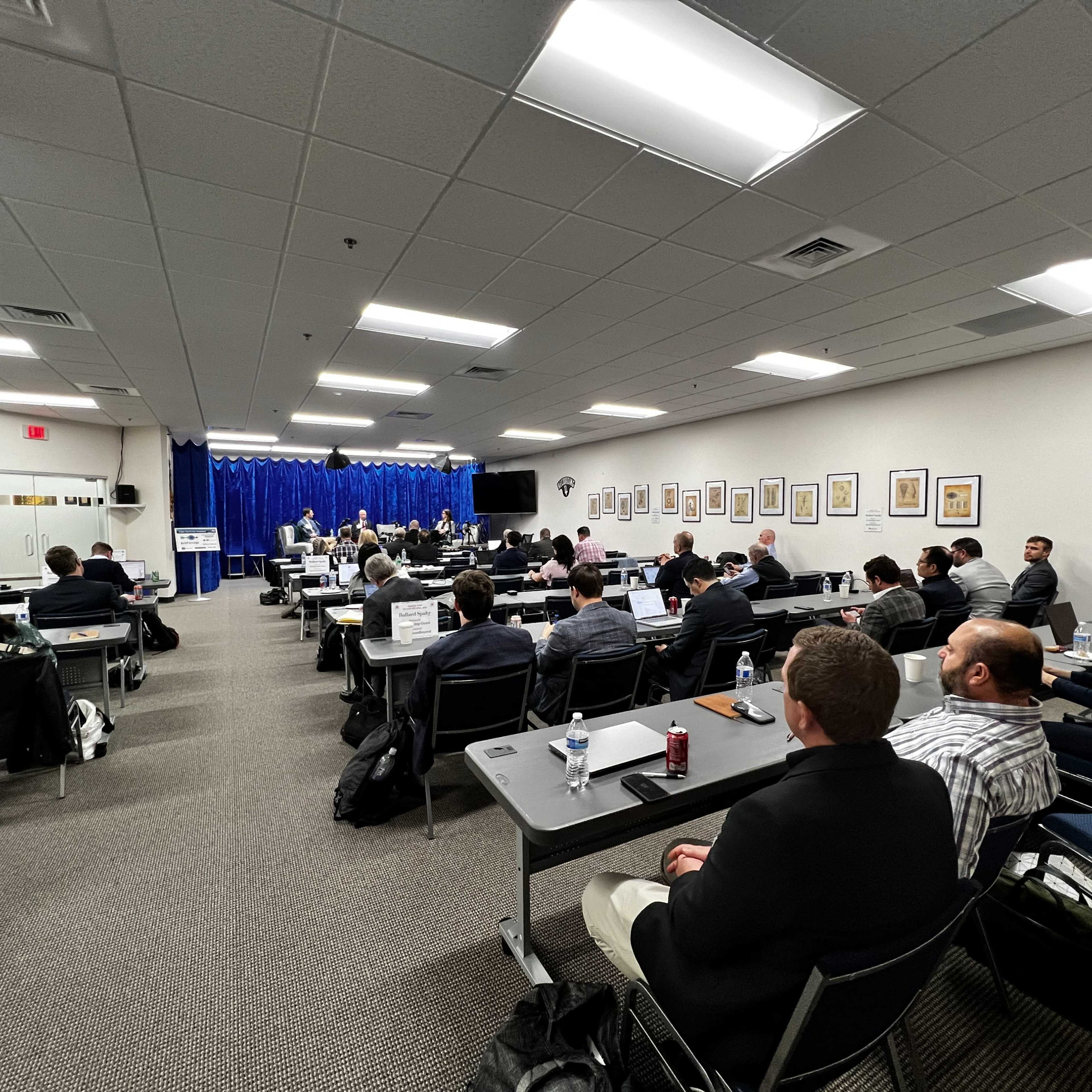 A group of attendees sits in a conference room facing a stage, where speakers are presenting in front of a blue curtain.
