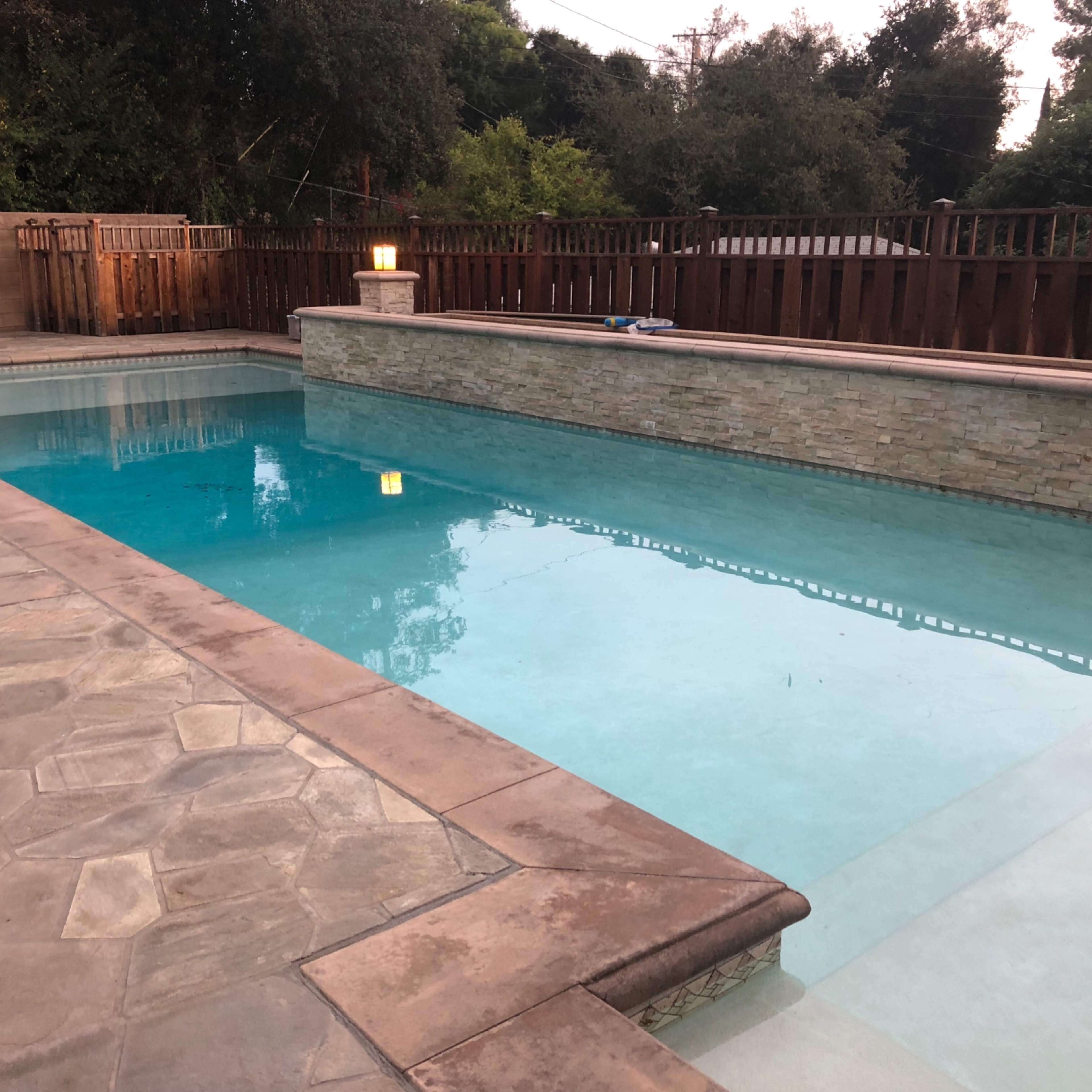 A rectangular swimming pool surrounded by a stone patio and wooden fencing, with trees visible in the background.