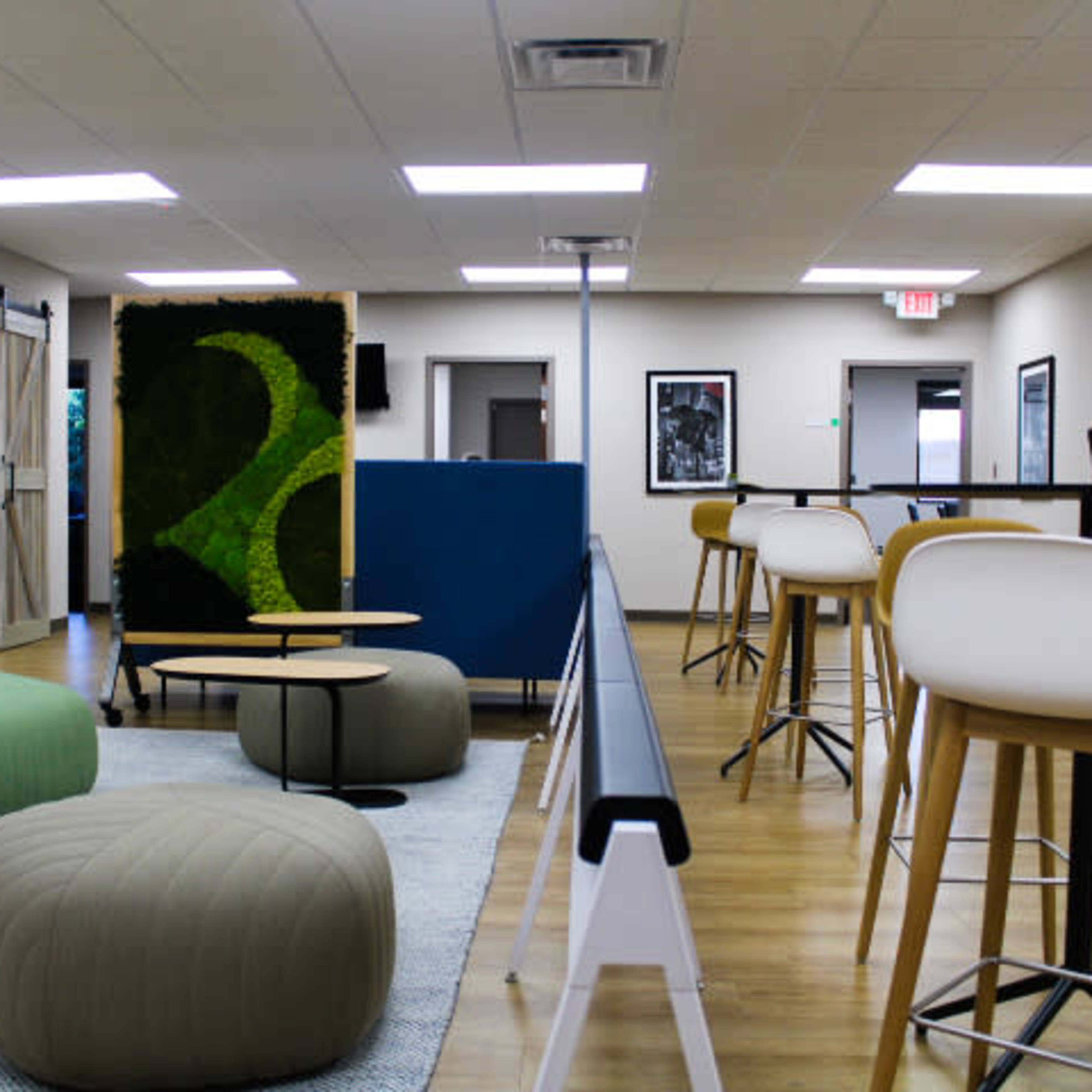 A modern office space with seating areas featuring round poufs and bar stools, surrounded by greenery and tasteful decor.