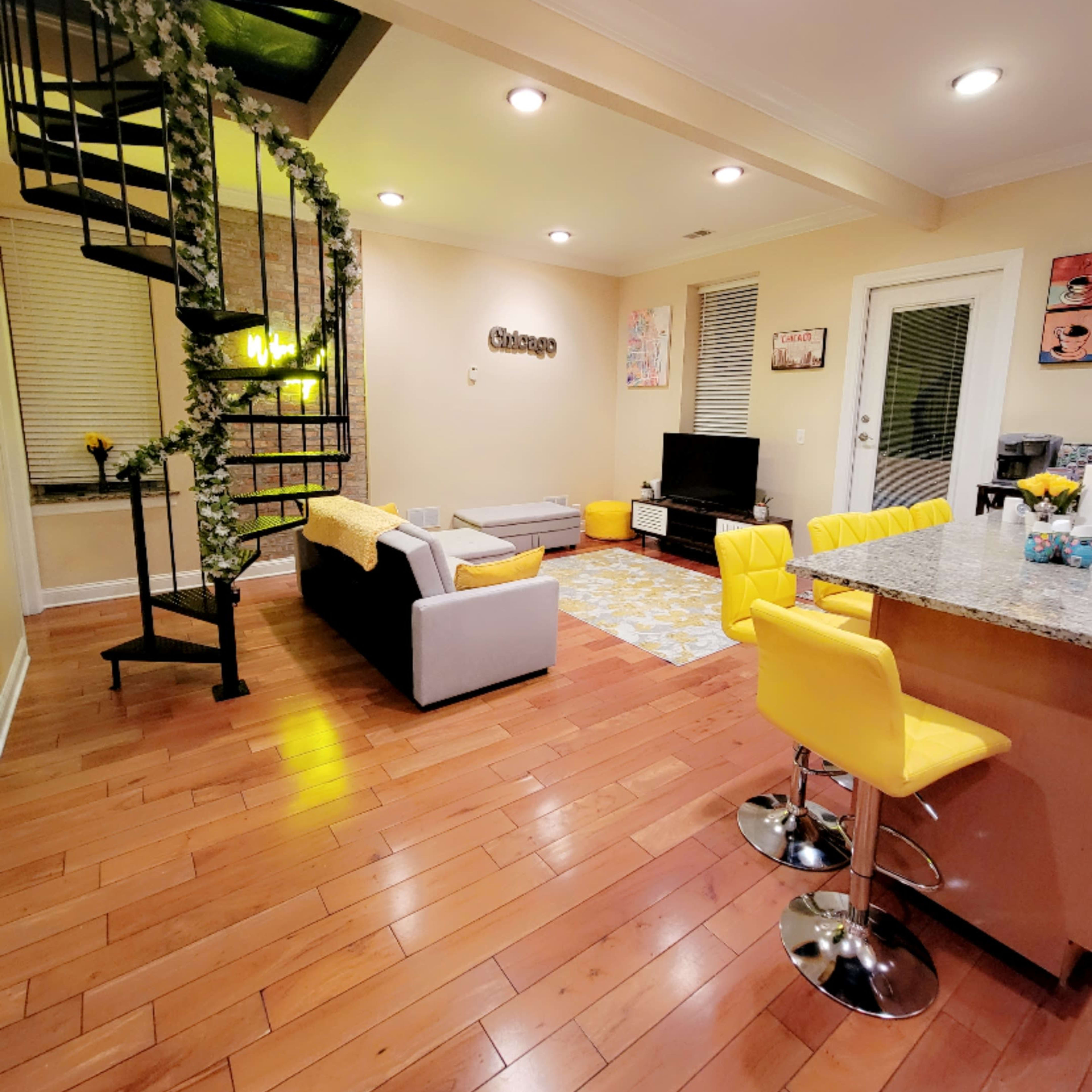 A modern living room and kitchen area with wooden flooring, a spiral staircase, yellow bar stools, and a gray sofa.