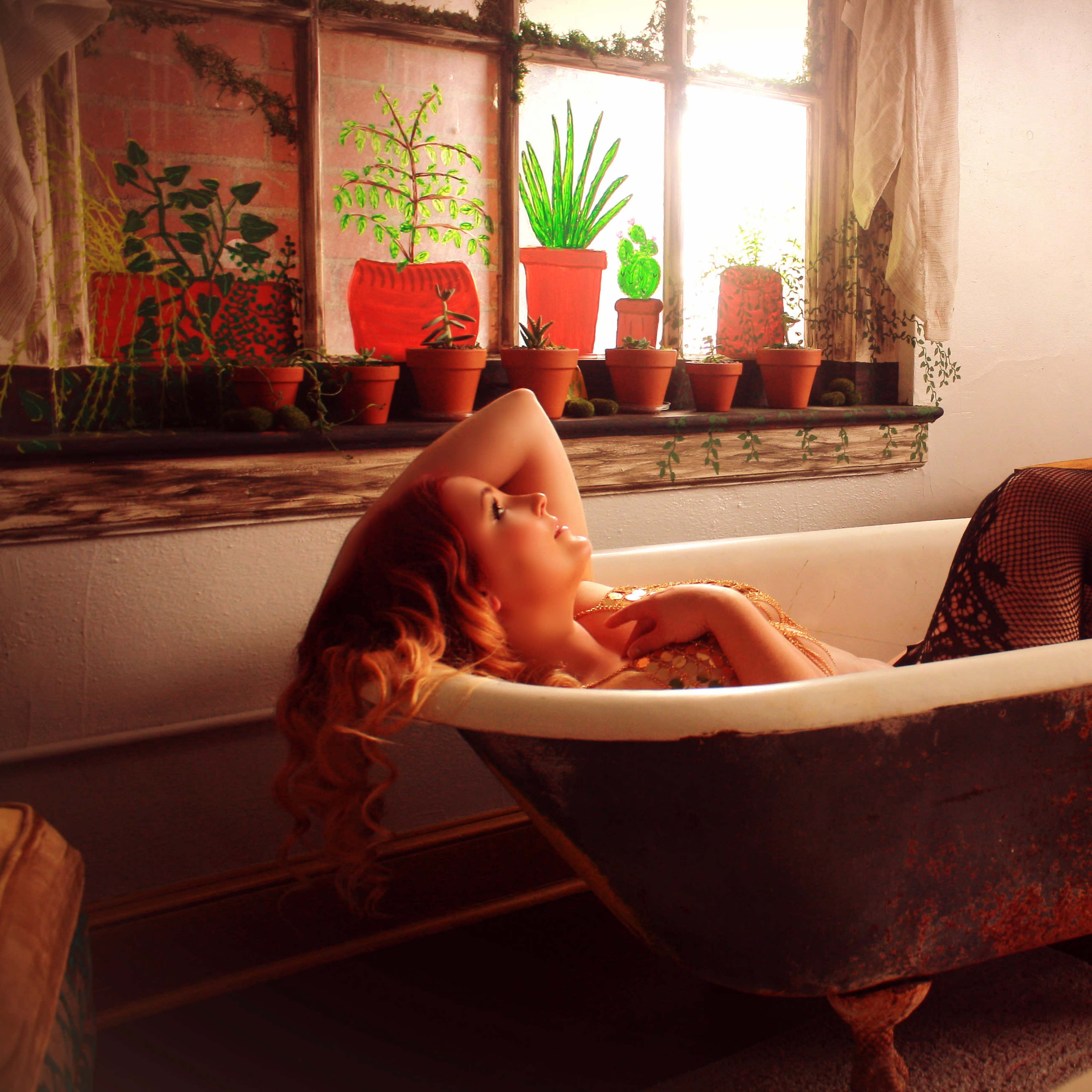 A woman with long hair lounges in a vintage bathtub surrounded by potted plants and natural light.