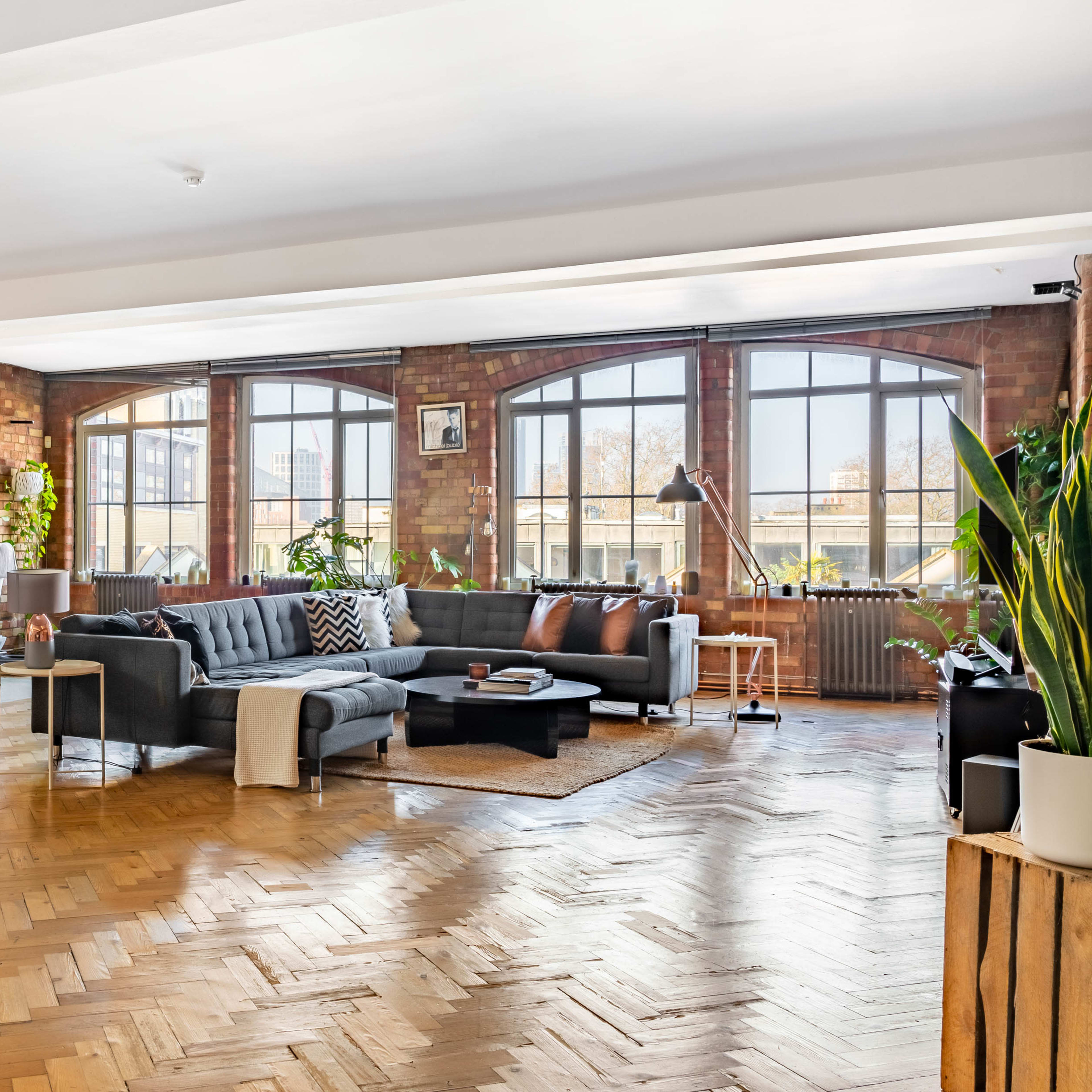 A spacious, modern living area with a gray sectional couch, wooden floors, and large windows allowing natural light to fill the room.