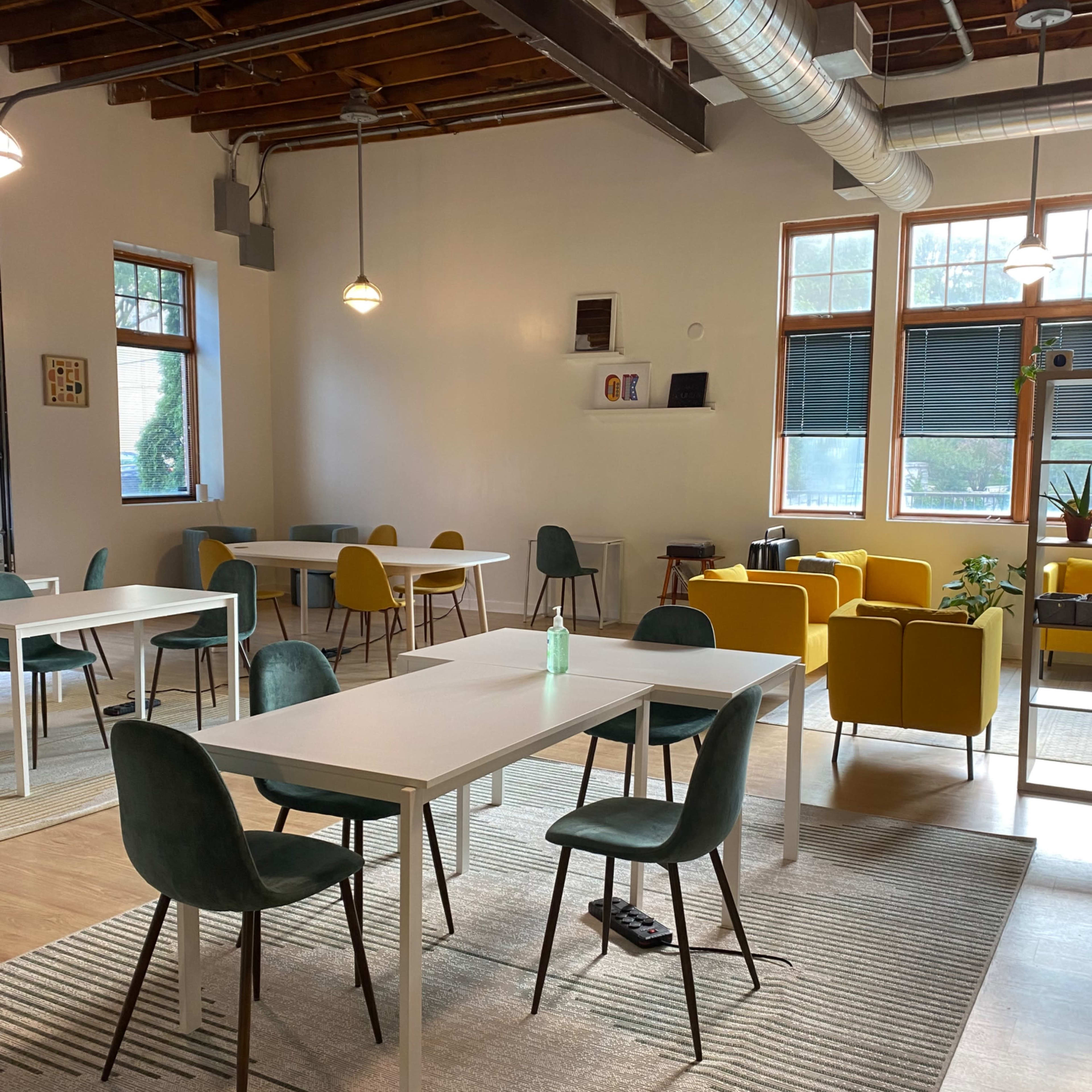 A brightly lit, modern workspace with several white tables surrounded by chairs in various colors, and a cozy seating area with yellow chairs.