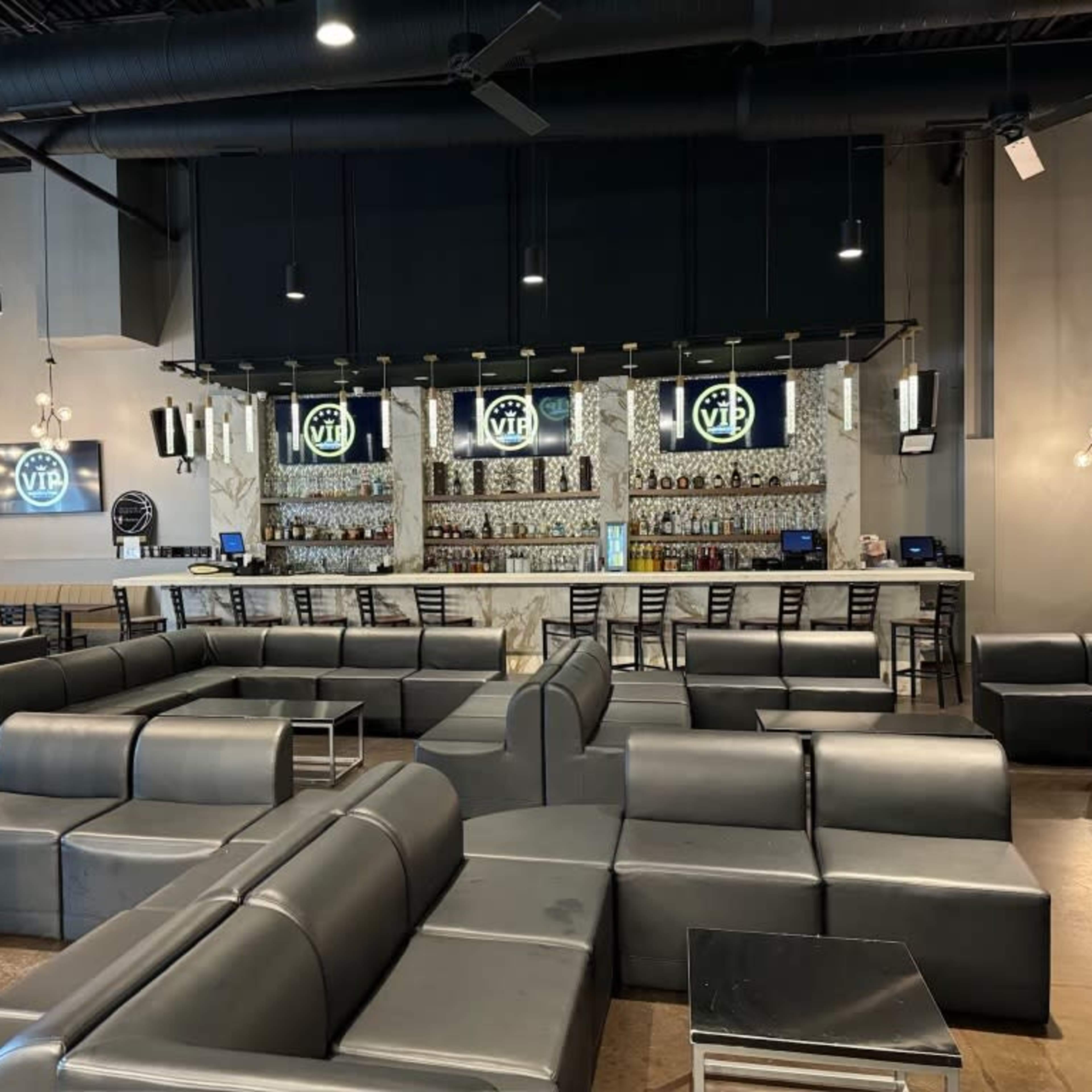 A spacious lounge area features black leather seating and a bar with illuminated shelves displaying various bottles.
