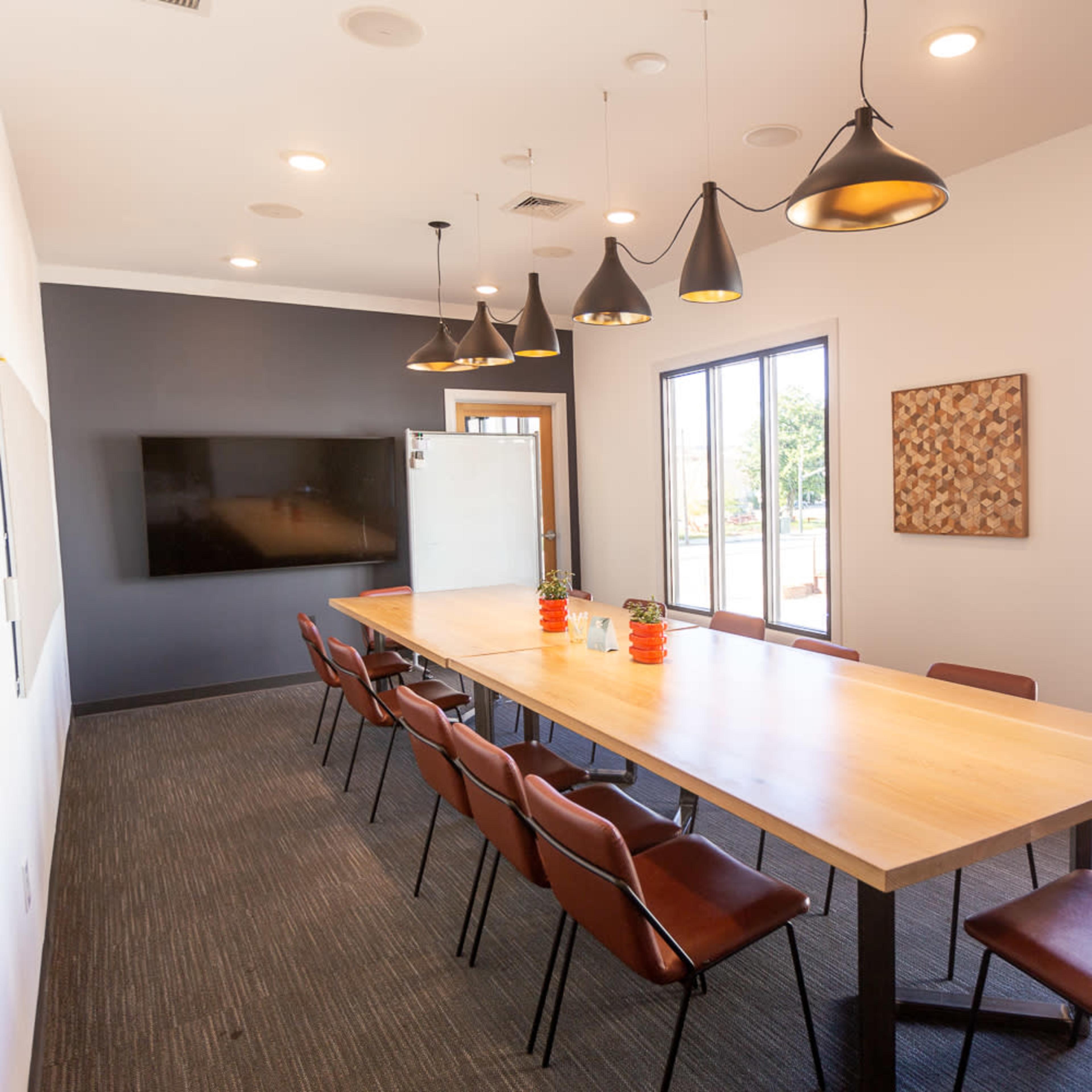 A modern conference room features a long wooden table surrounded by dark chairs, with pendant lights overhead and large windows letting in natural light.