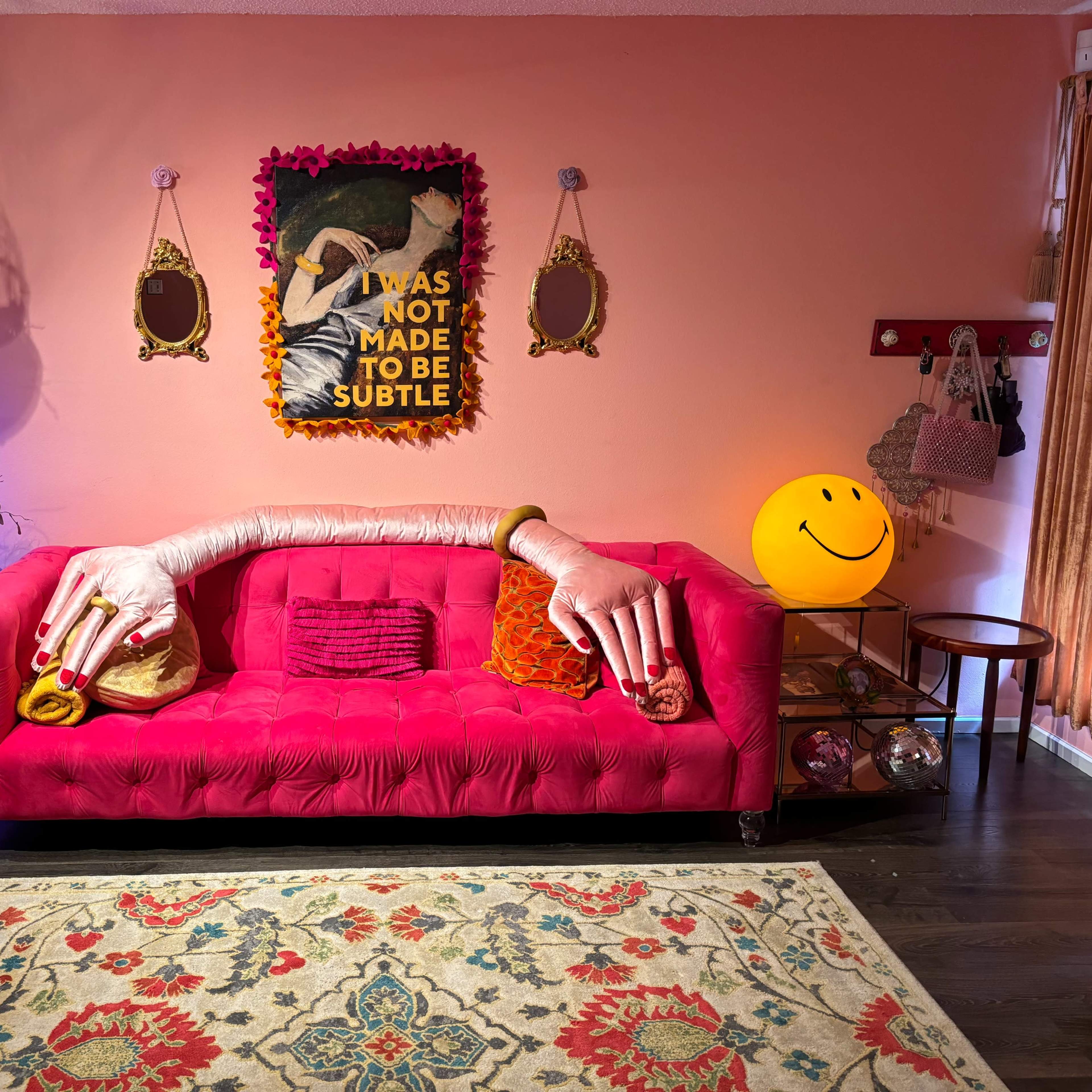 A vibrant pink couch with oversized decorative hands rests against a peach wall, surrounded by eclectic decor, including mirrors, a smiling lamp, and a colorful rug.