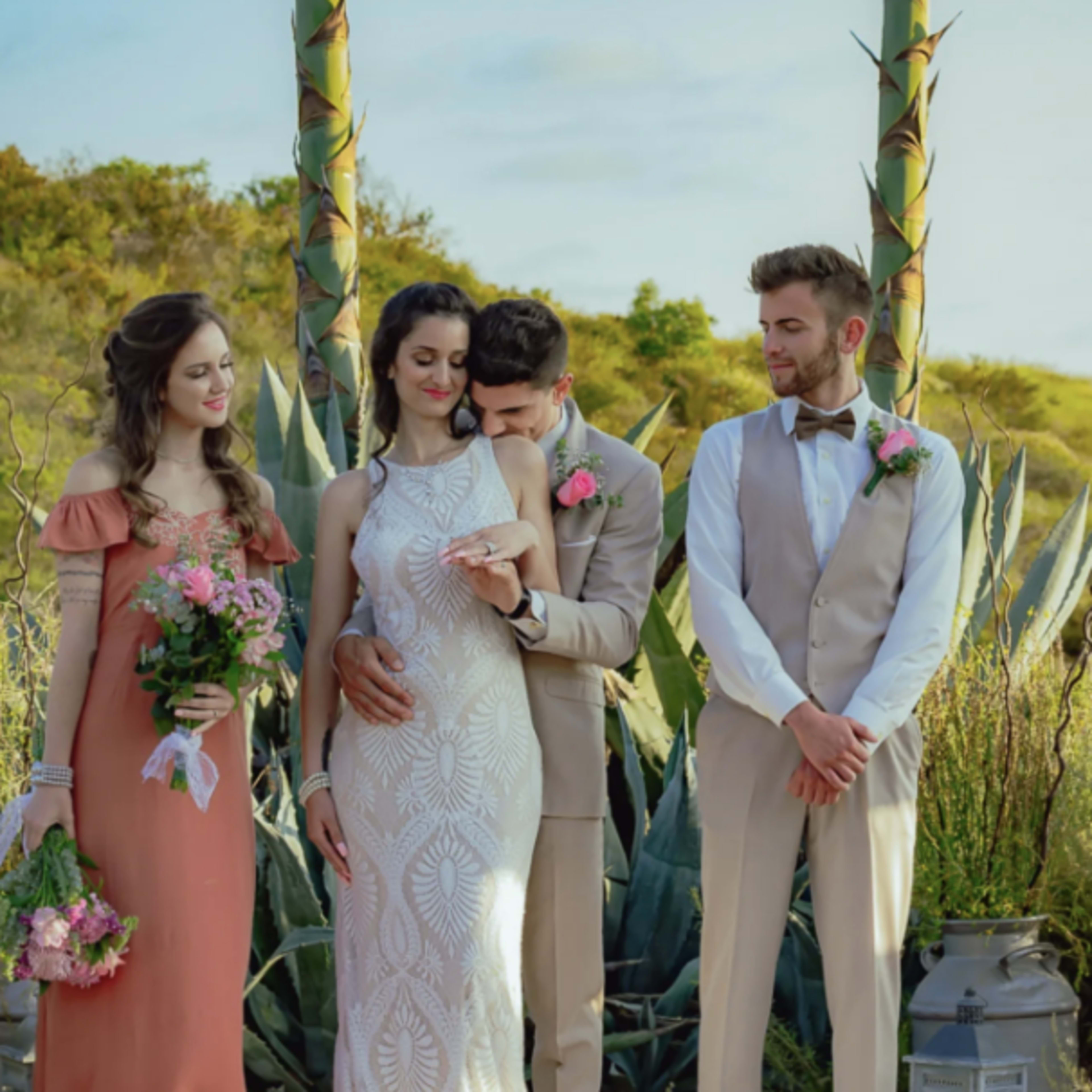 A couple stands at the altar during a wedding ceremony, flanked by their attendants, with a backdrop of large agave plants and decorative lanterns.