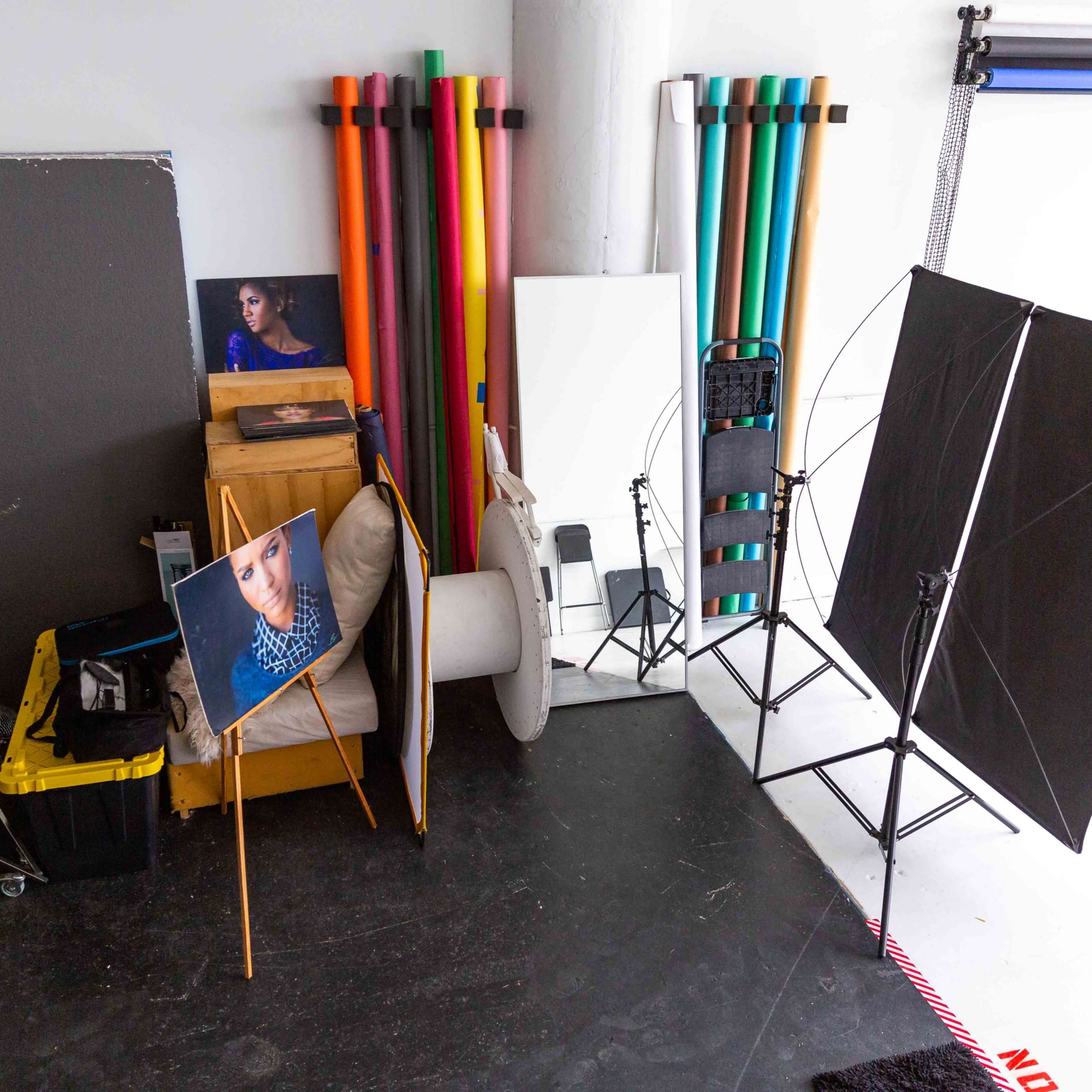 A photography studio with a corner setup featuring a portrait on an easel, backdrop rolls, lighting equipment, and various studio props.