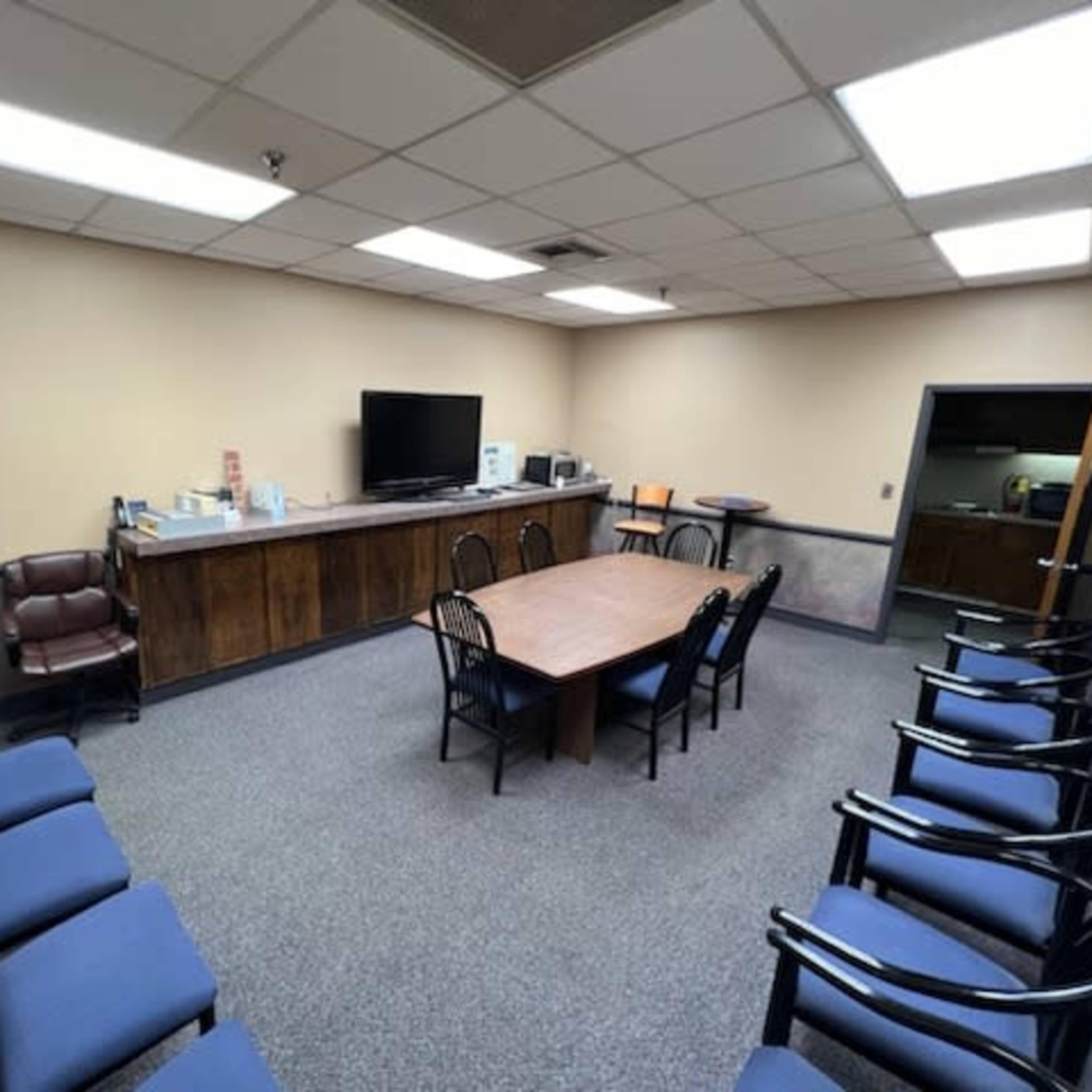A waiting room with a long table and chairs, a television, and a counter with a printer and office supplies.