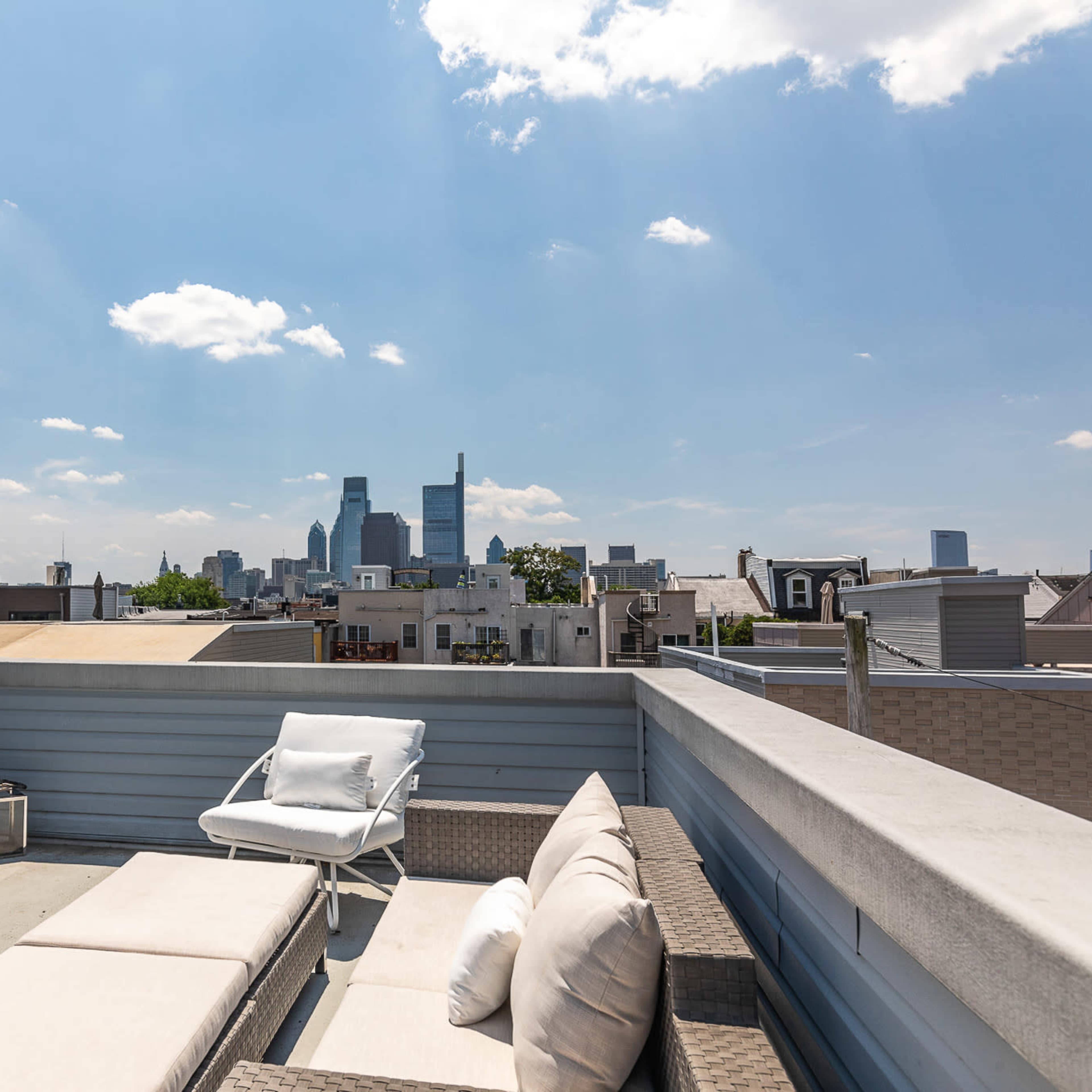 A rooftop terrace features lounge chairs overlooking a city skyline under a partly cloudy sky.