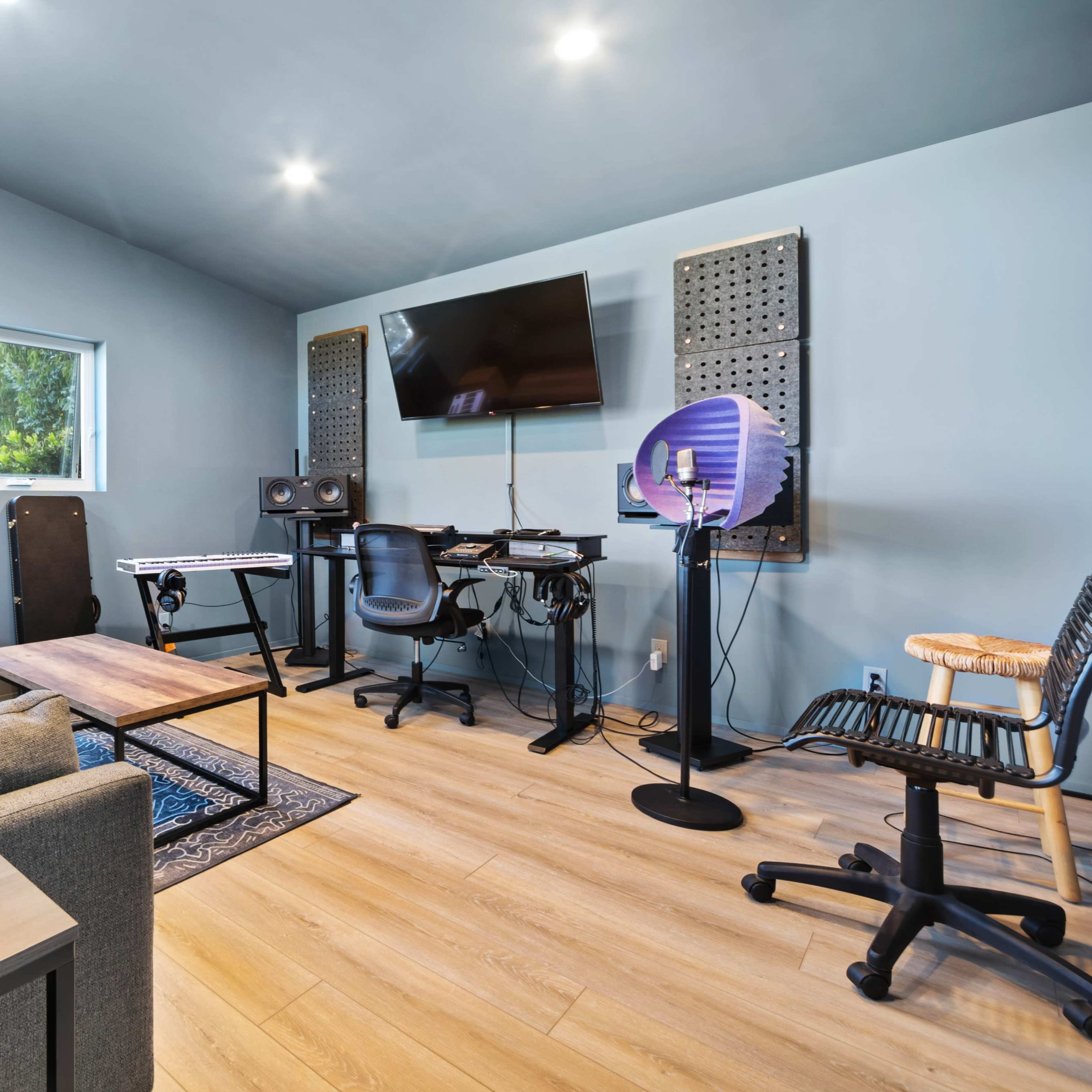 A music recording studio features a gray sofa, a wooden table, a desk with audio equipment, and soundproof panels on the walls.