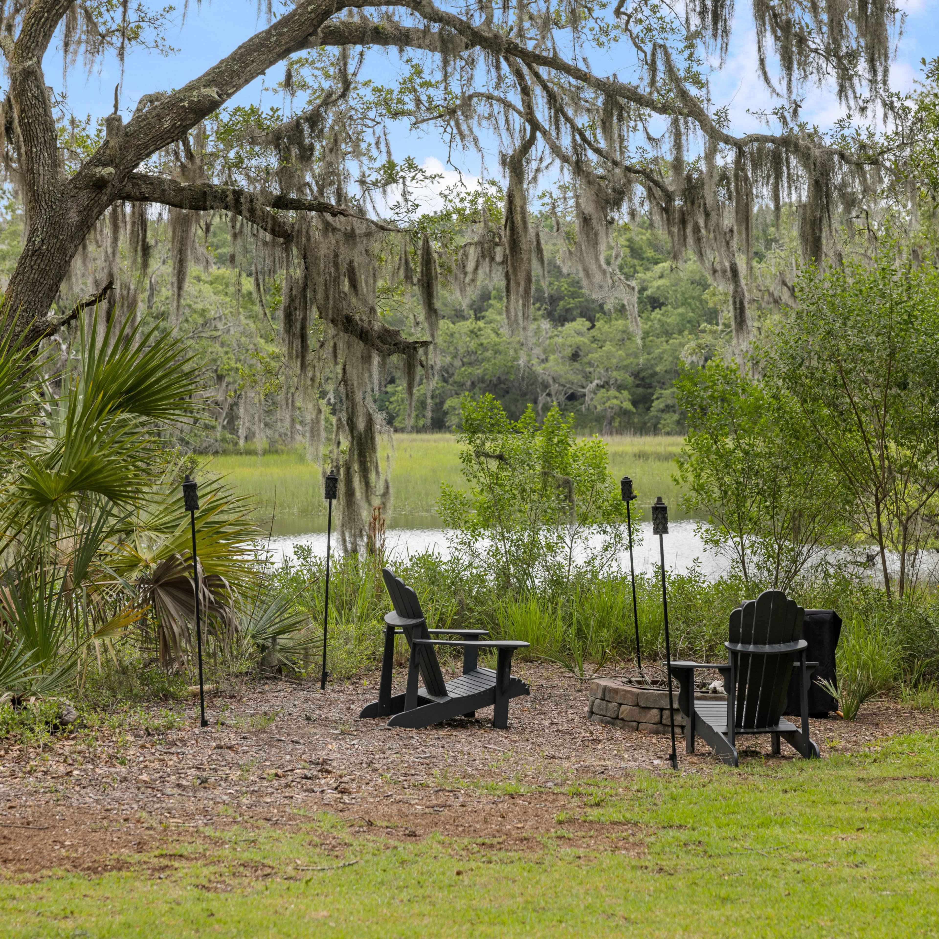 The scene features two black Adirondack chairs around a stone fire pit, surrounded by lush greenery and moss-draped trees near a calm body of water.