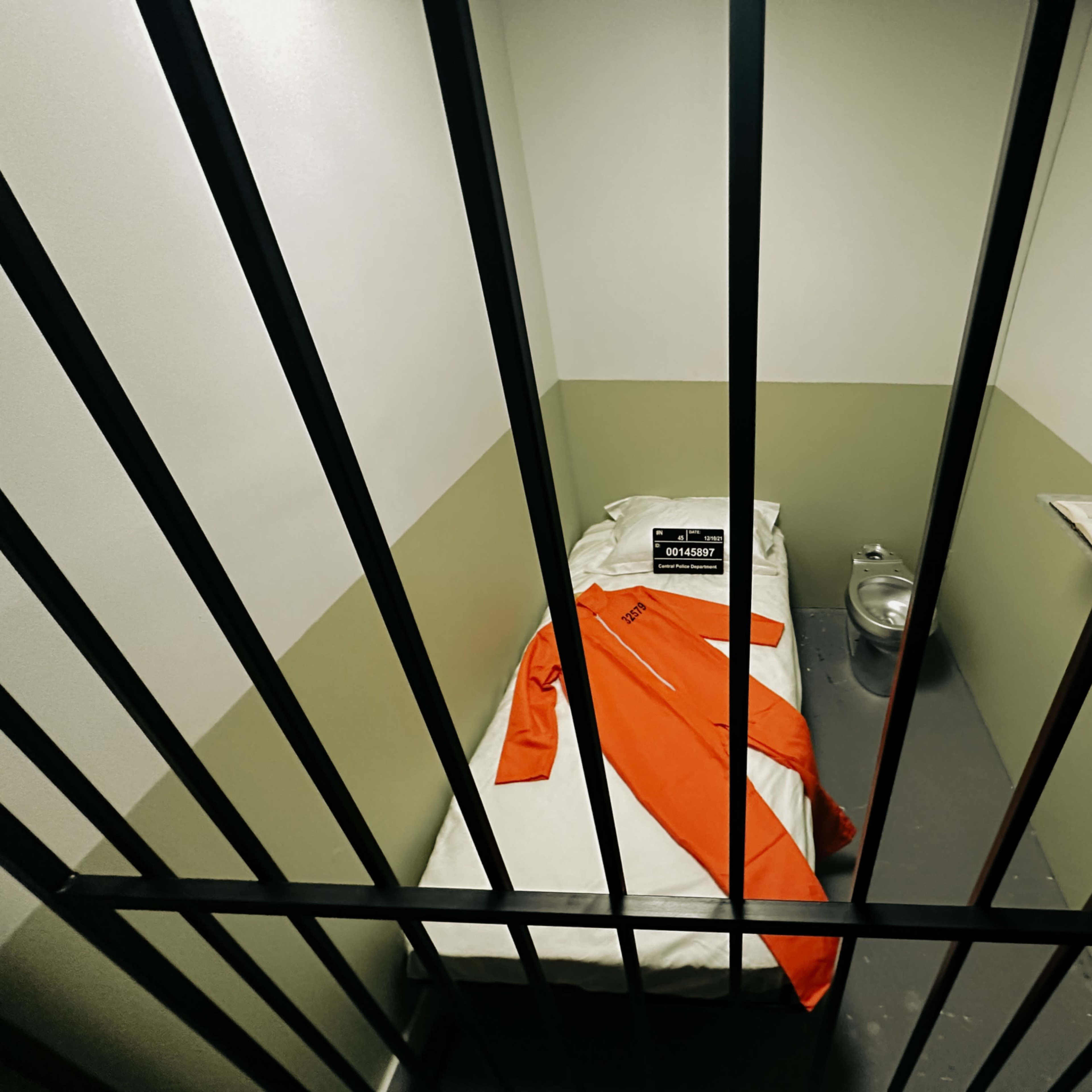 A small jail cell features a bed with white linens, an orange jumpsuit laid across it, a metal toilet, and a window with bars.