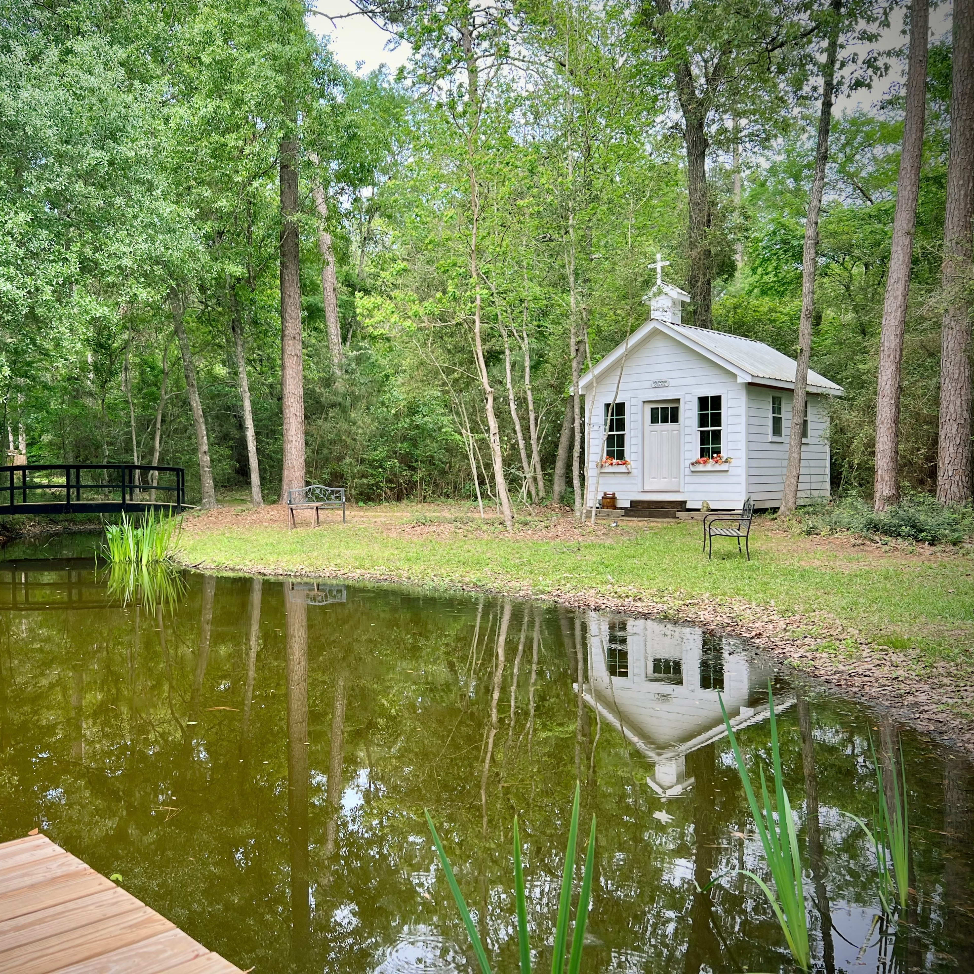 A small, white cabin with flower boxes stands by a pond surrounded by trees, with a wooden bridge visible in the background.