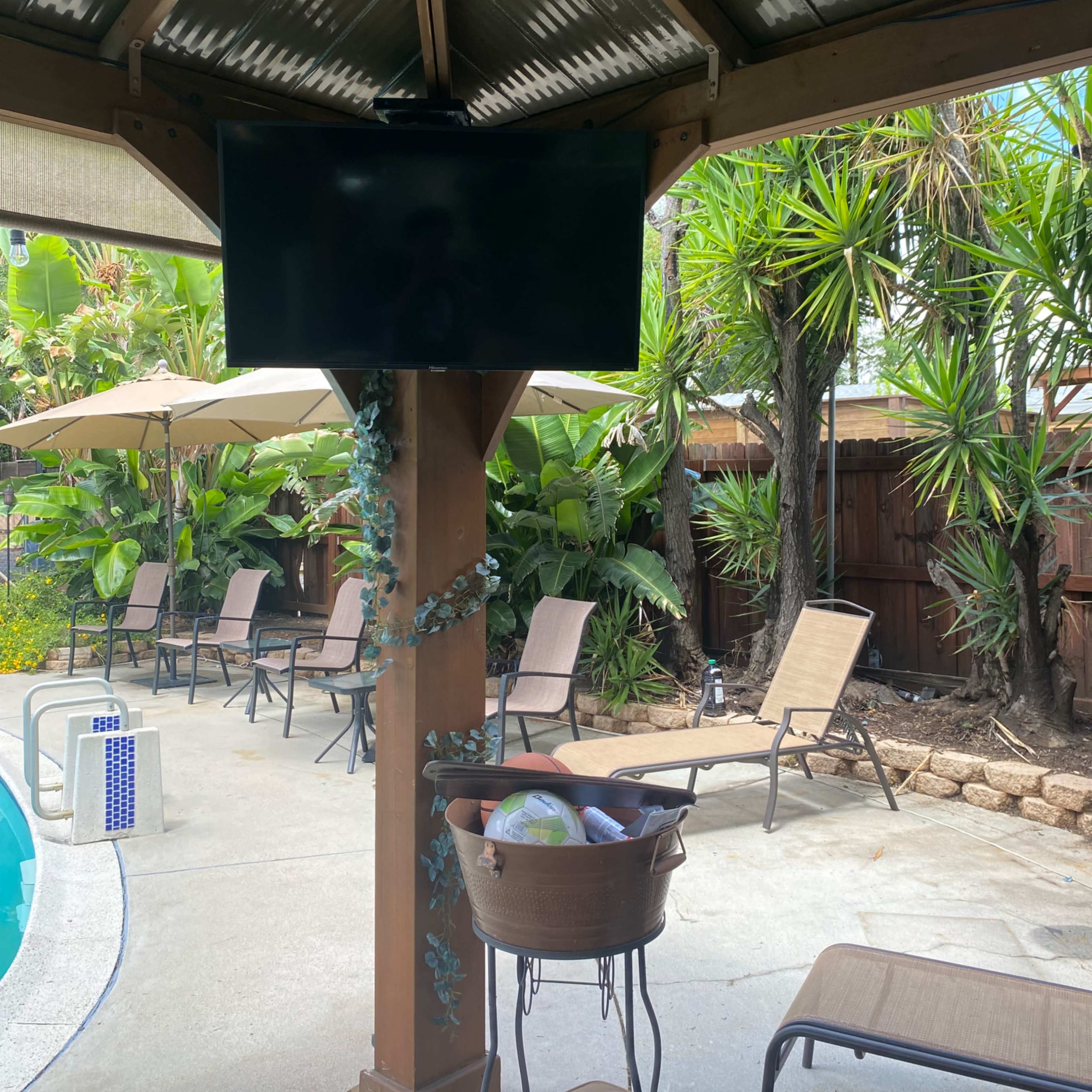 A covered outdoor area with a TV mounted on a post, surrounded by lounge chairs and tropical plants near a swimming pool.