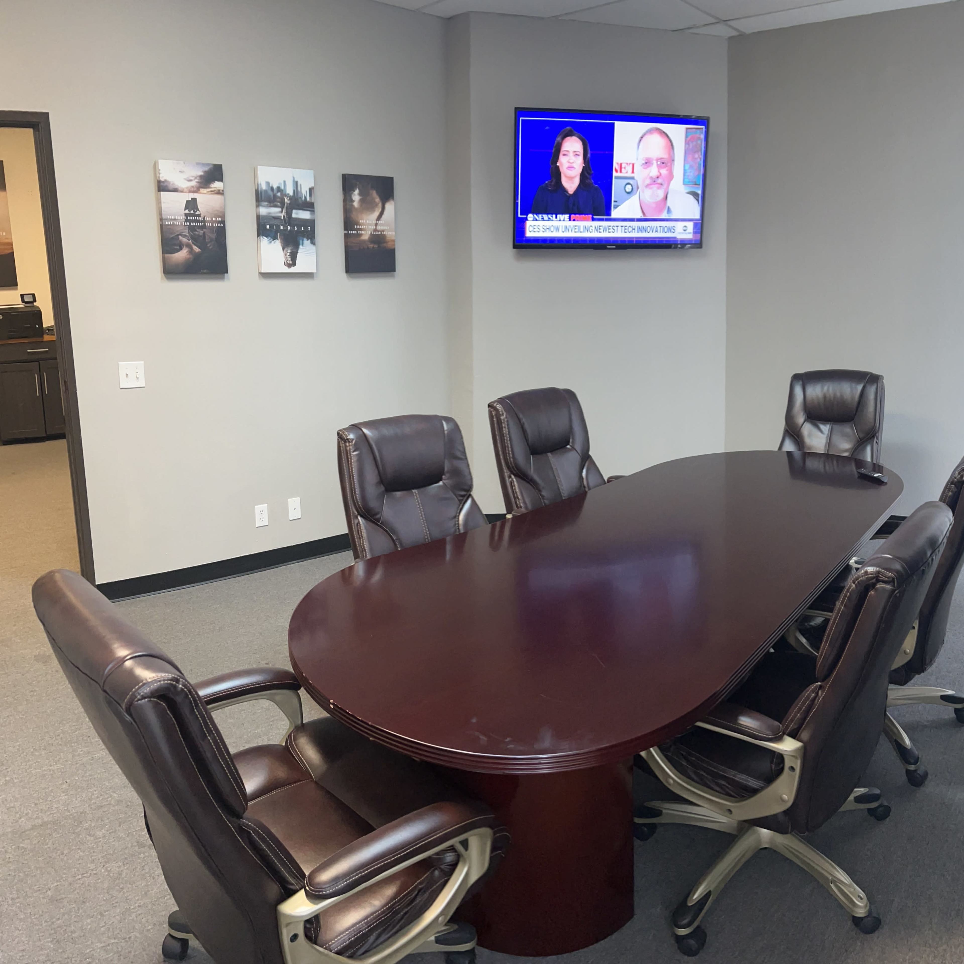 A conference room featuring a large oval table surrounded by six leather chairs, with a television displaying a news broadcast on the wall.