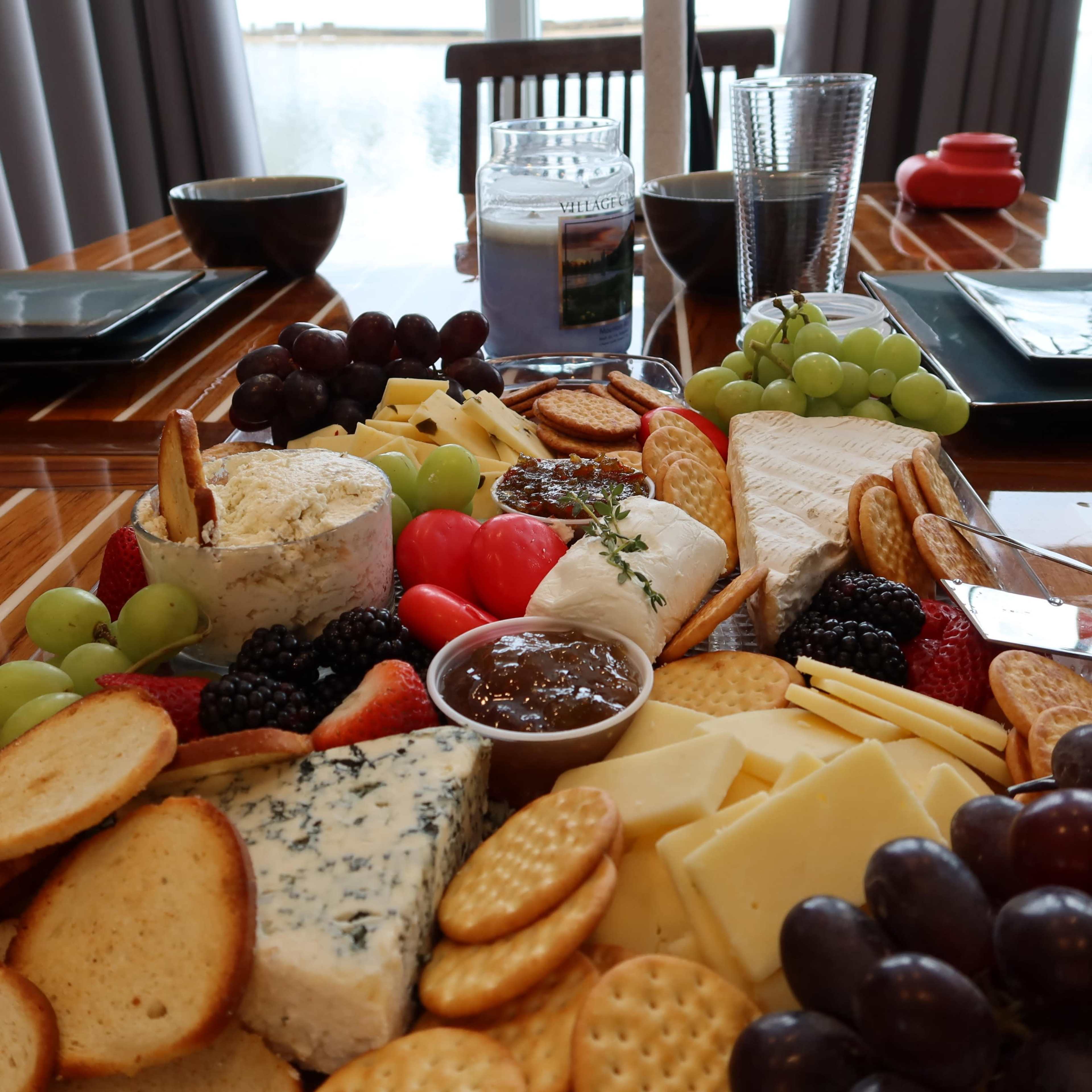 A wooden table is set with a large cheese and fruit platter featuring a variety of cheeses, crackers, fruits, and spreads, alongside glassware and decorative bowls.