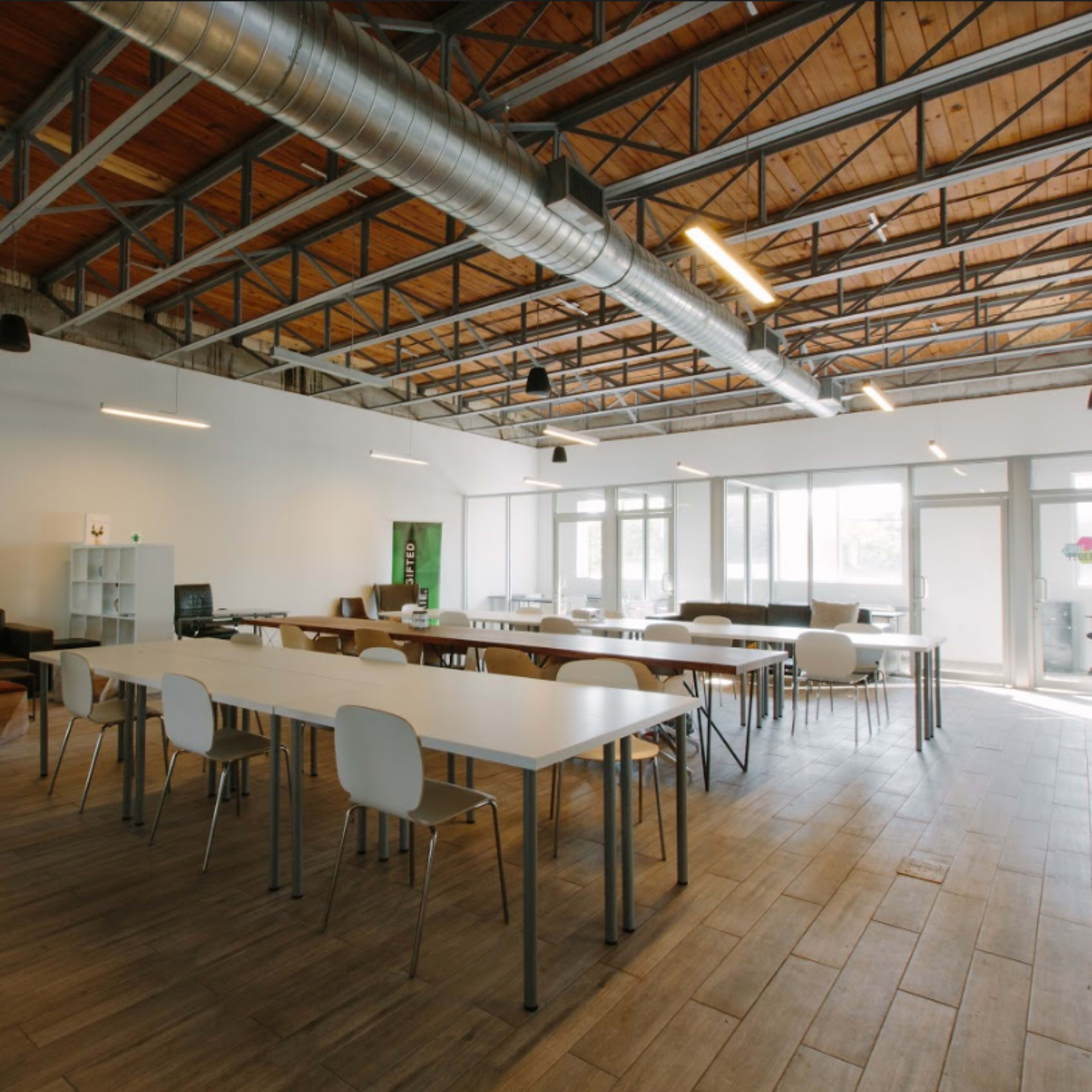 A spacious, modern office interior with long white tables and chairs, large windows allowing natural light, and exposed wooden beams.