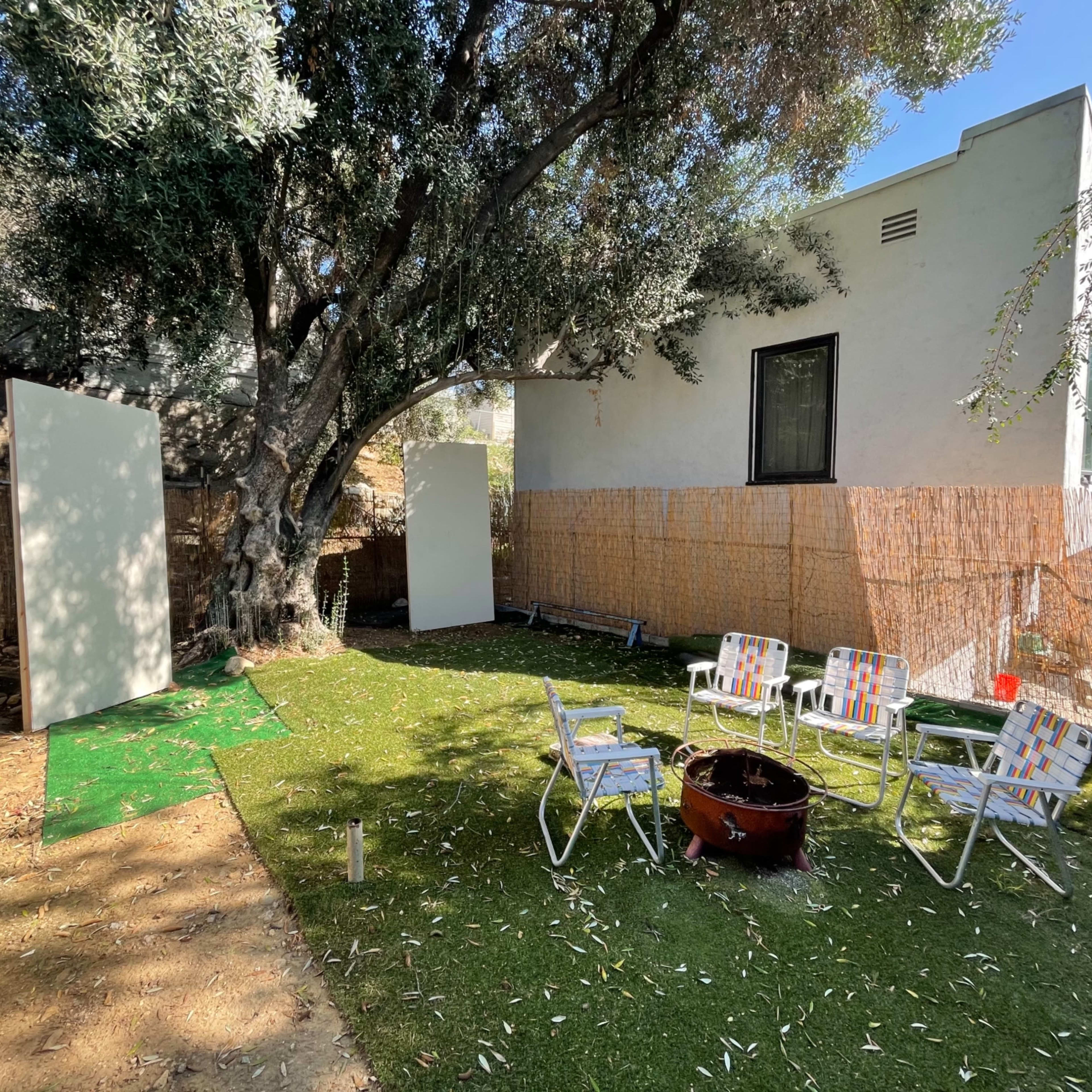 A backyard with green artificial grass, a fire pit in the center, and two white panels set up against a backdrop of bamboo fencing and trees.