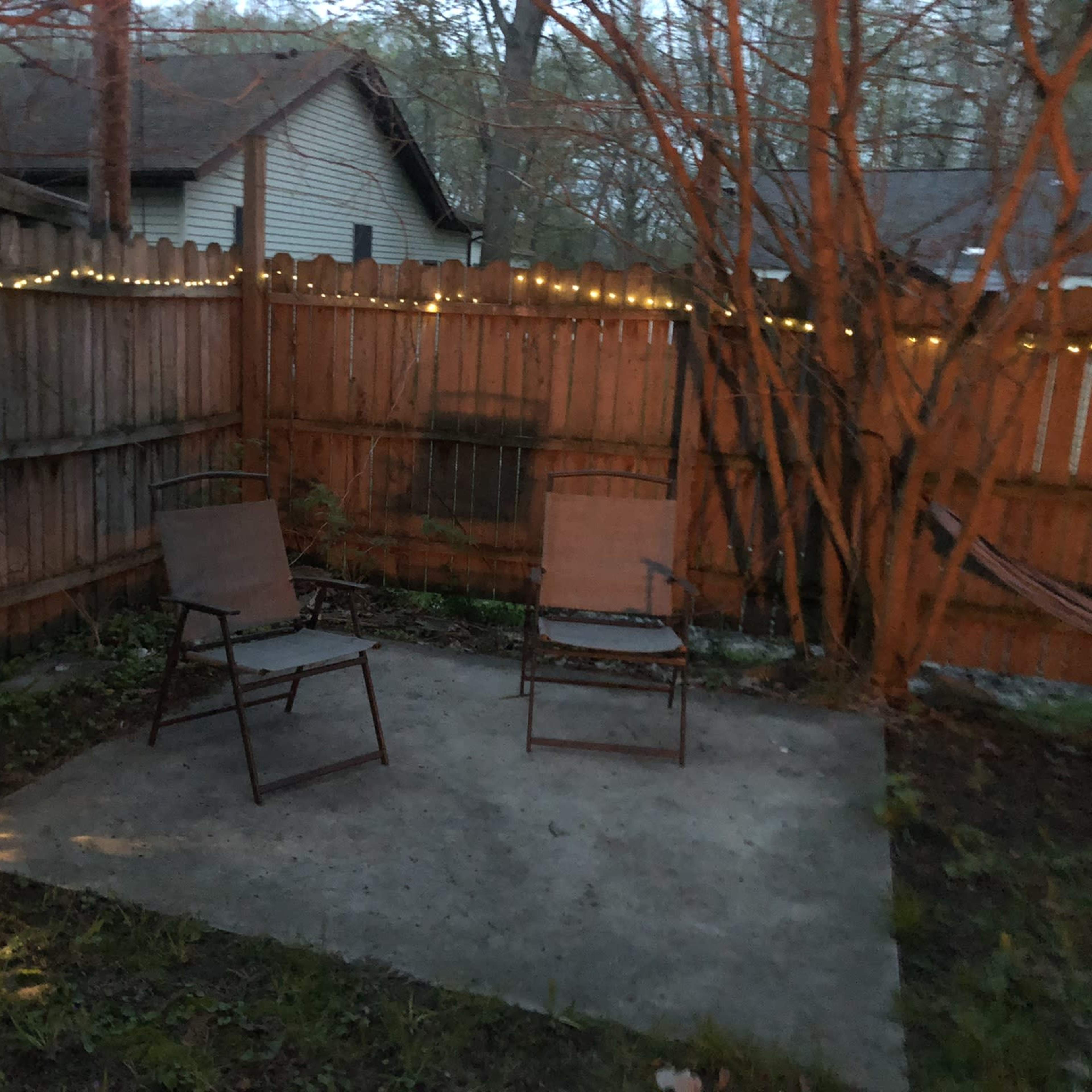 A small outdoor patio area with two chairs on a concrete slab, surrounded by a wooden fence and illuminated by string lights.