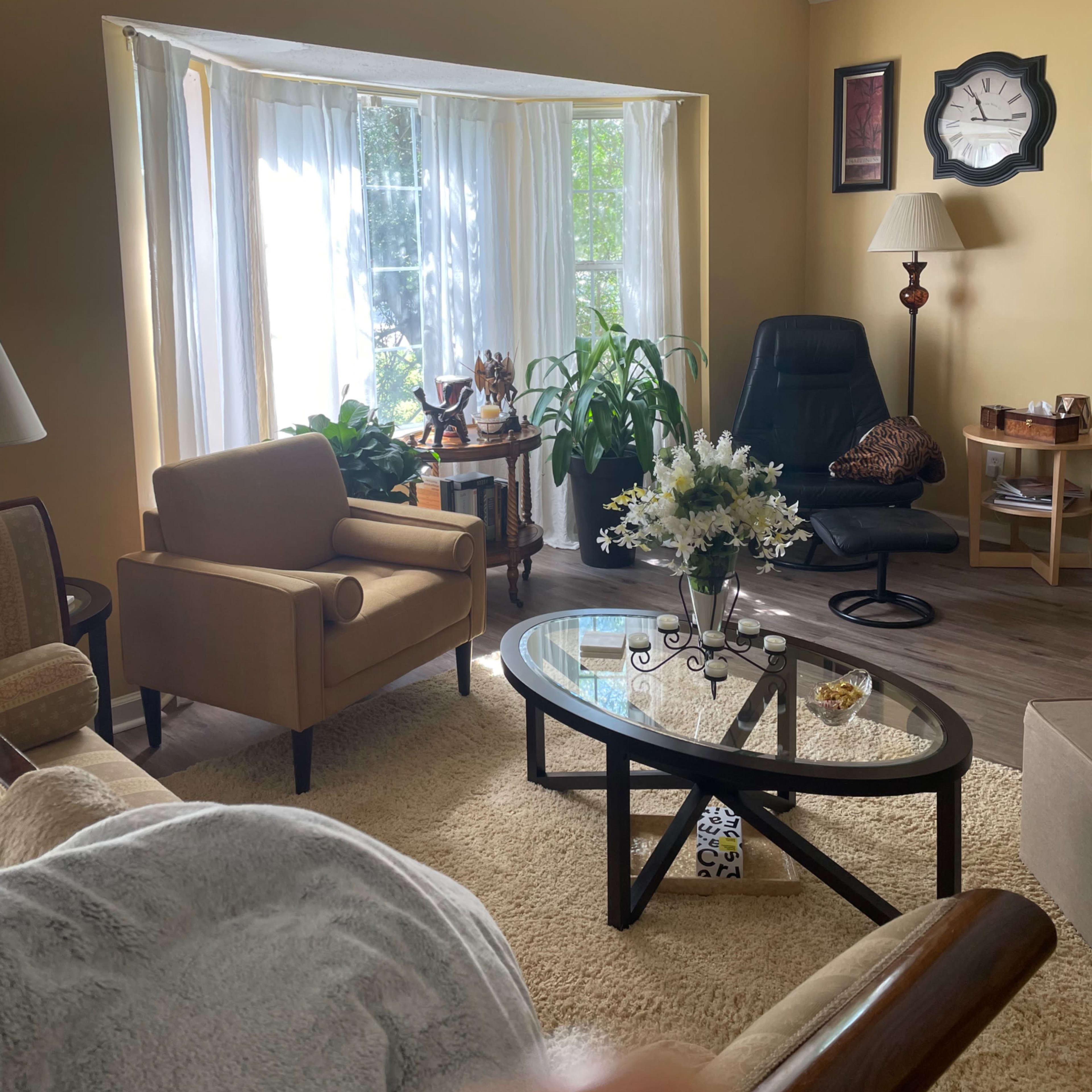 A cozy living room features a large window with white curtains, two armchairs, a coffee table with flowers, and a corner lamp.