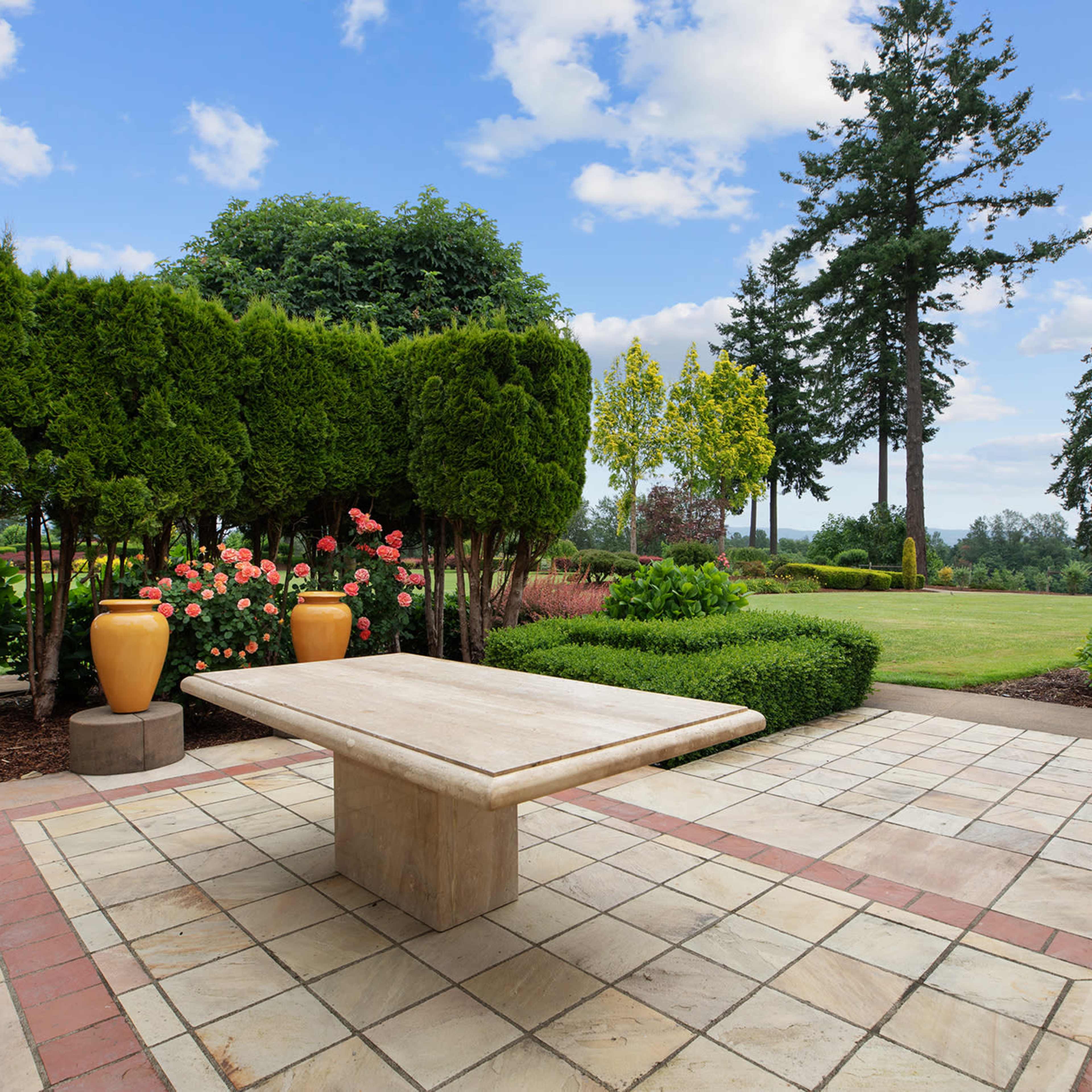 A landscaped outdoor area features a stone table surrounded by greenery and floral arrangements under a blue sky.