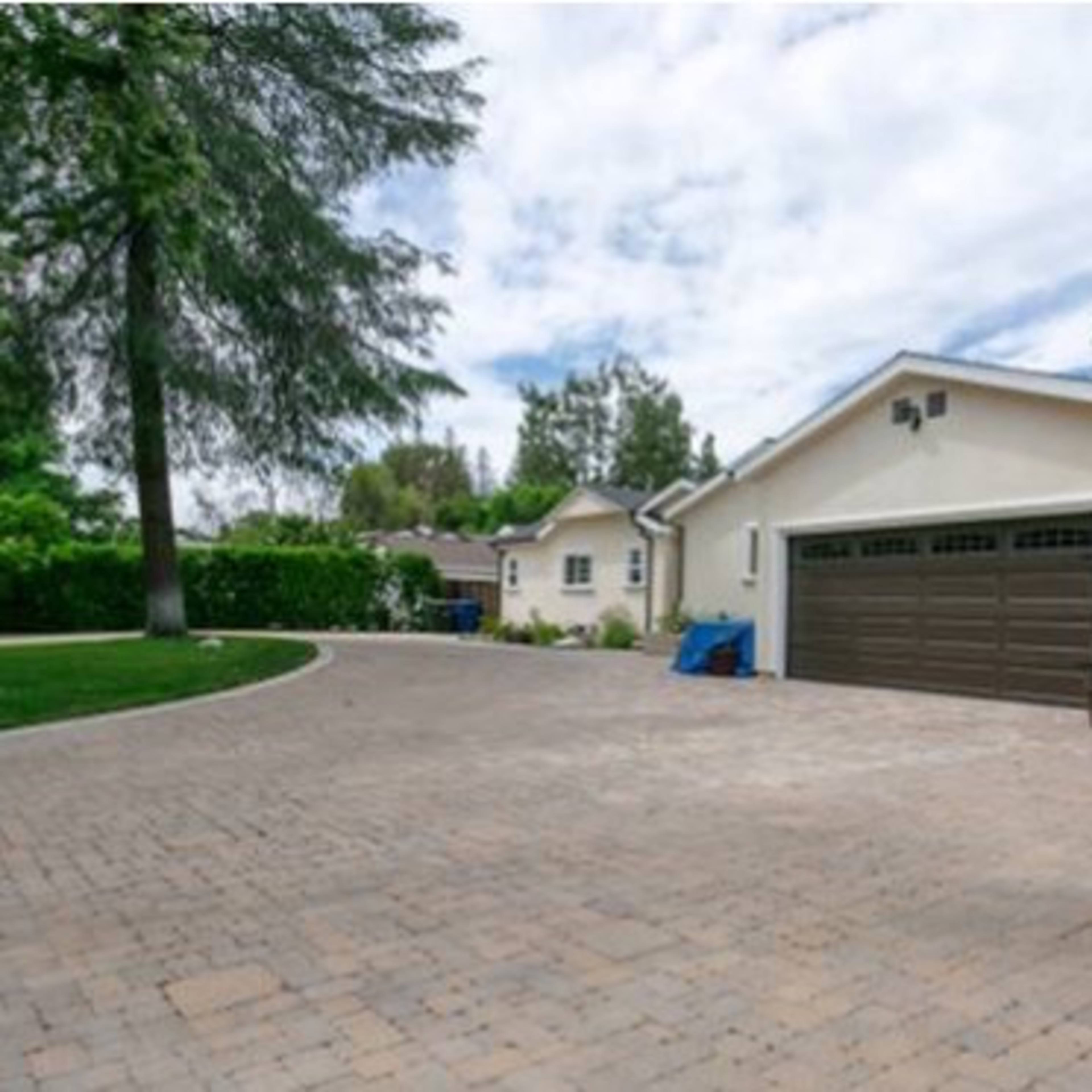 A brick-paved driveway leads to a beige house with a dark garage door, surrounded by green trees and hedges.