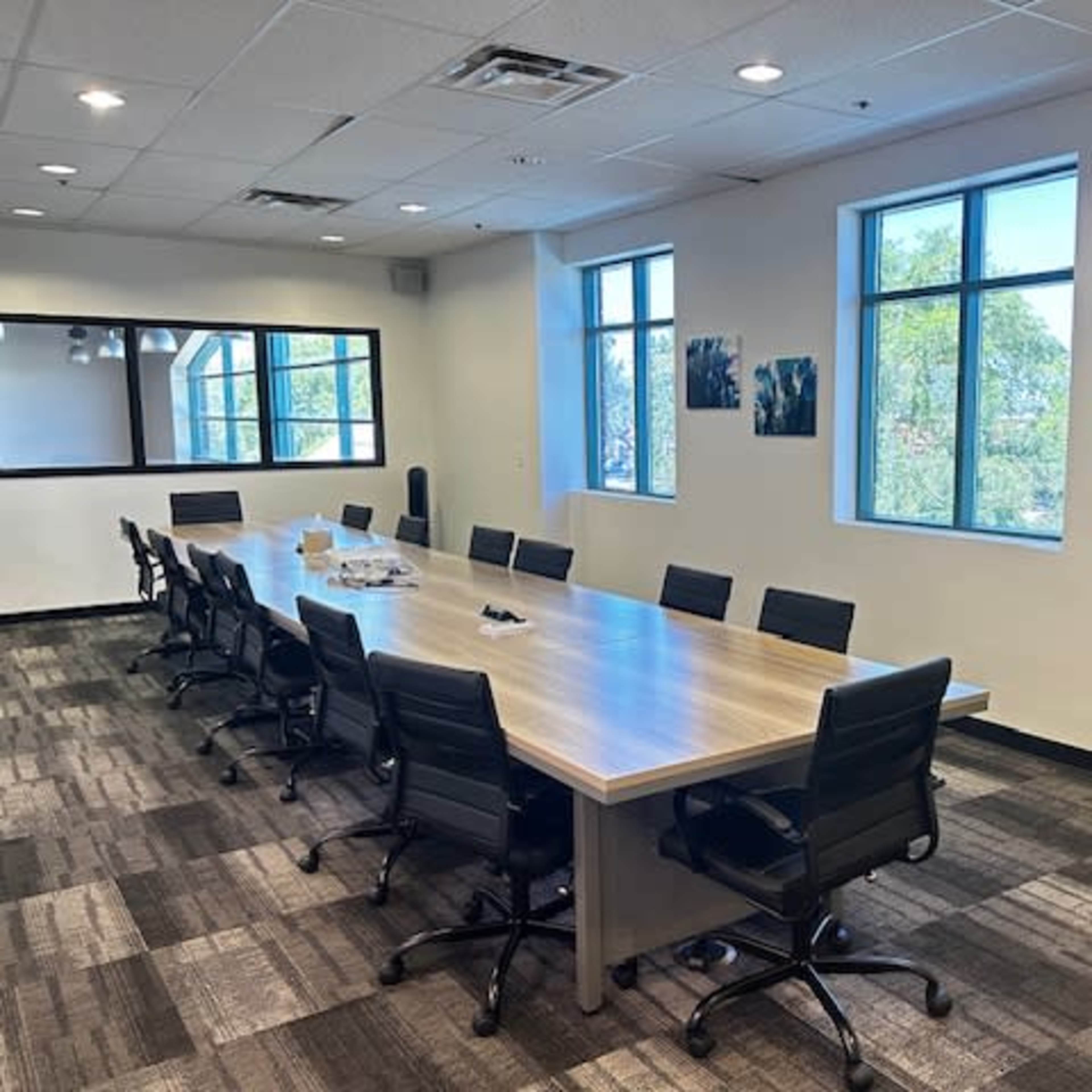 A large conference room features a long wooden table surrounded by black chairs, with windows providing natural light and artwork on the walls.