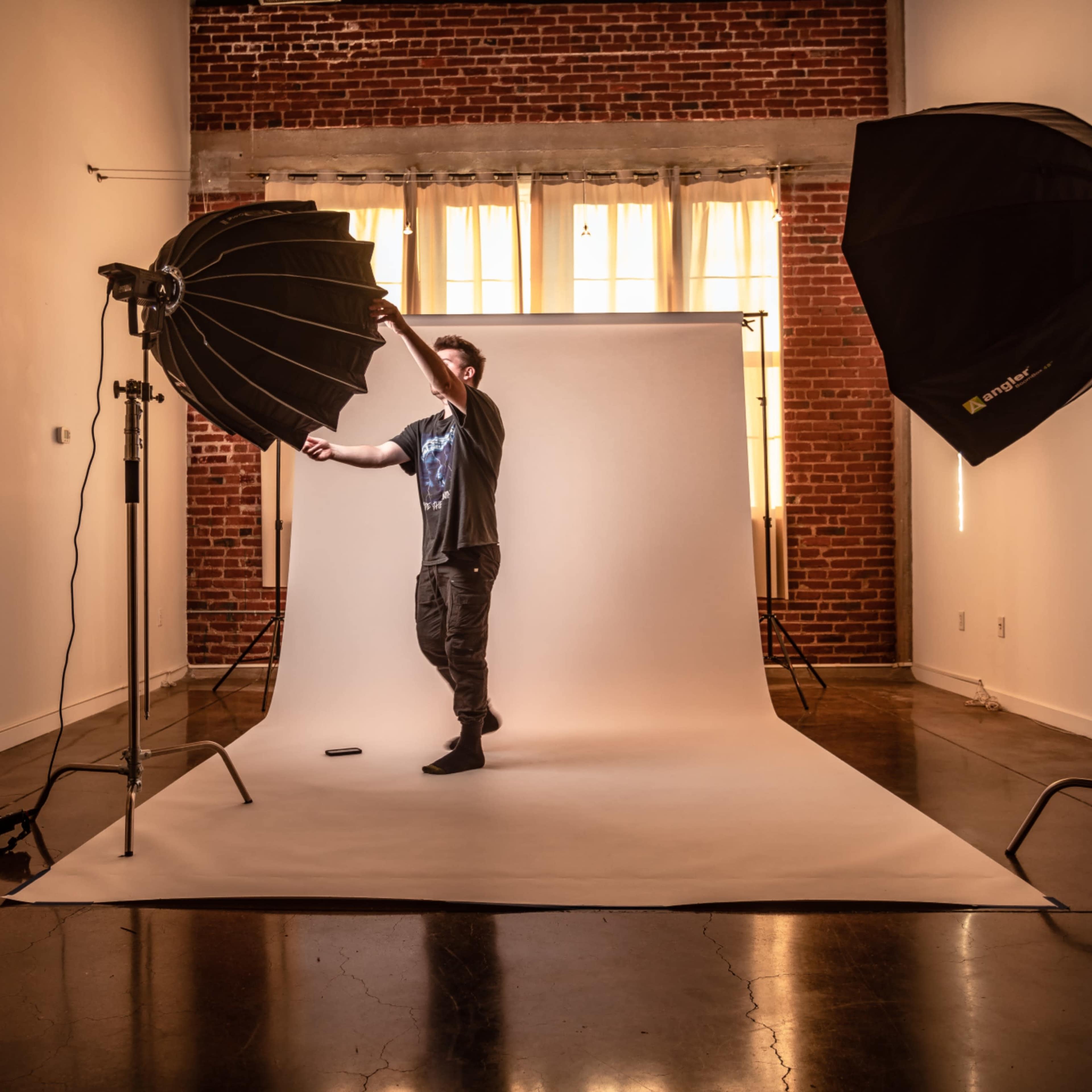 A photographer adjusts a large softbox light in a studio featuring a minimalist backdrop and exposed brick walls.