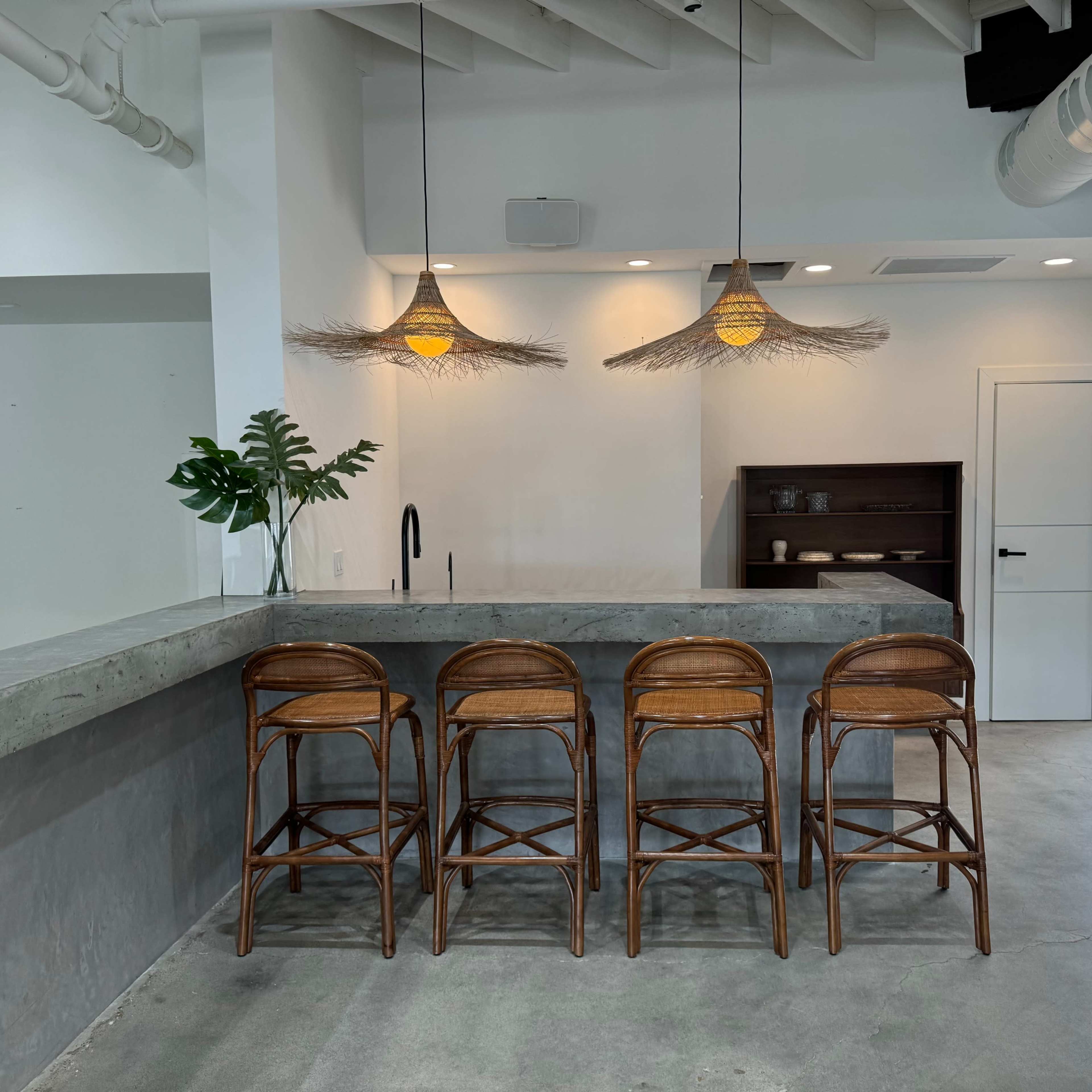 A modern kitchen features a concrete bar with four rattan stools, illuminated by pendant lights, and a small plant on the counter.