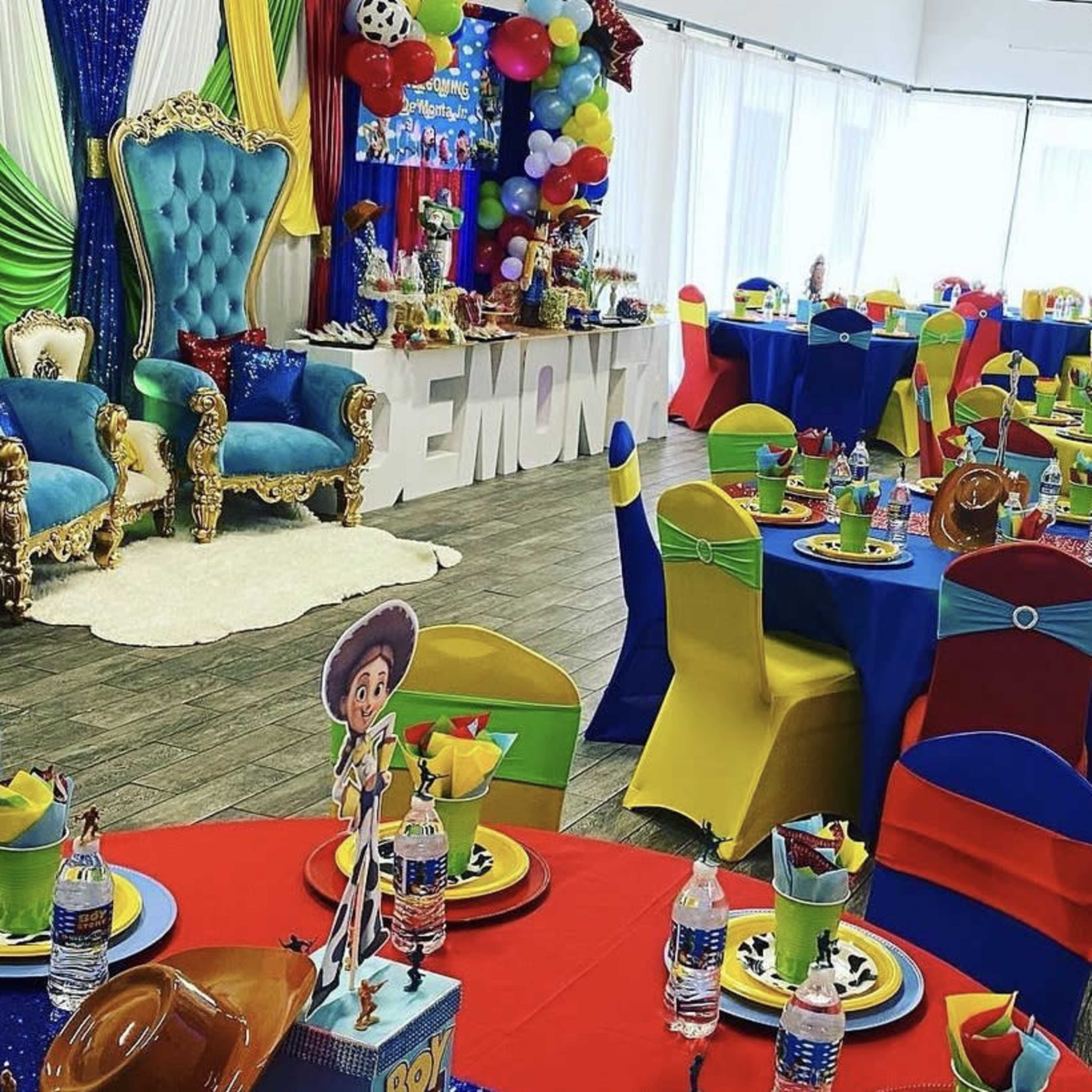 A colorful party venue decorated with blue and gold chairs, vibrant tablecloths in red, blue, yellow, and green, and festive balloons.