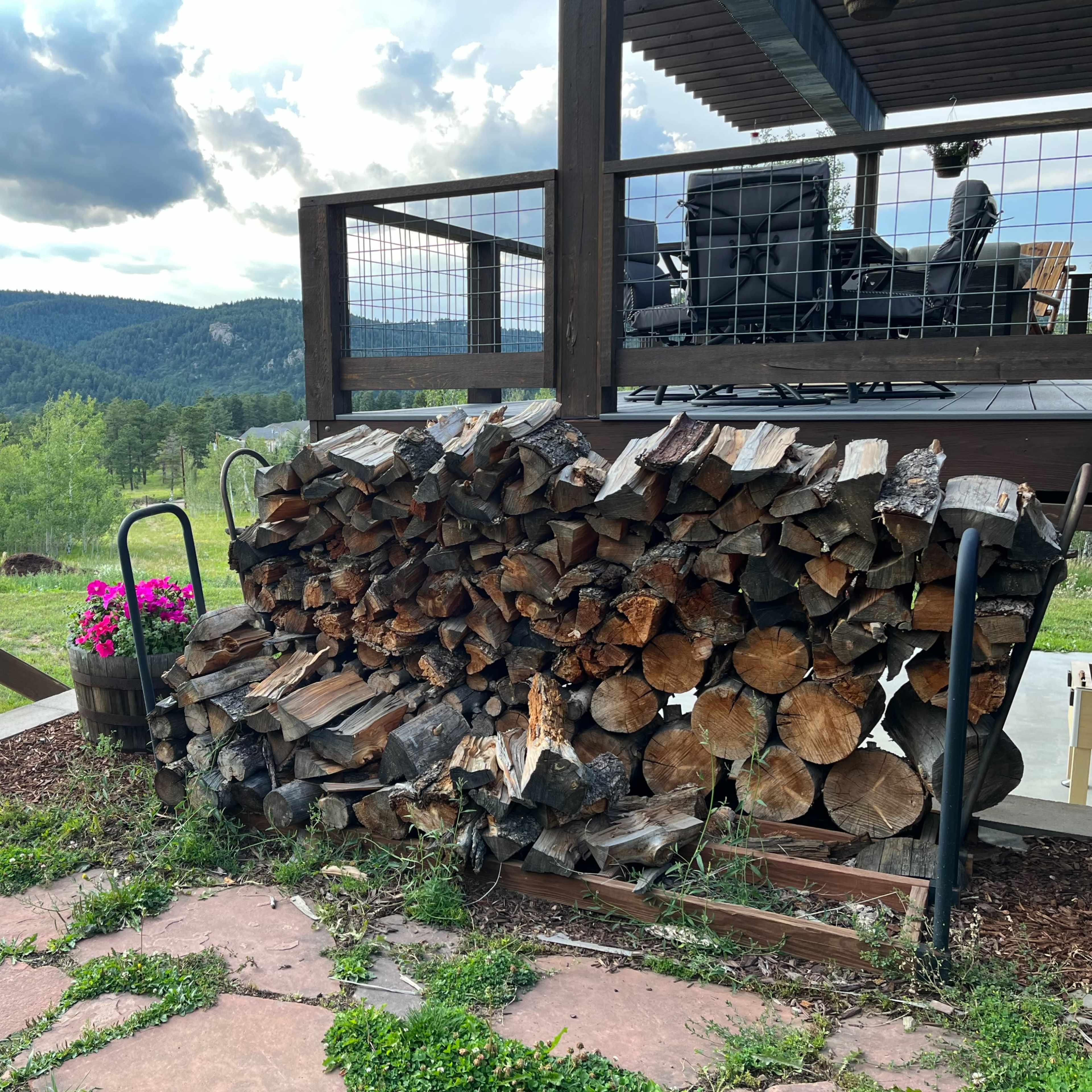 A stack of firewood rests beside a wooden deck with chairs overlooking a green landscape and distant mountains.