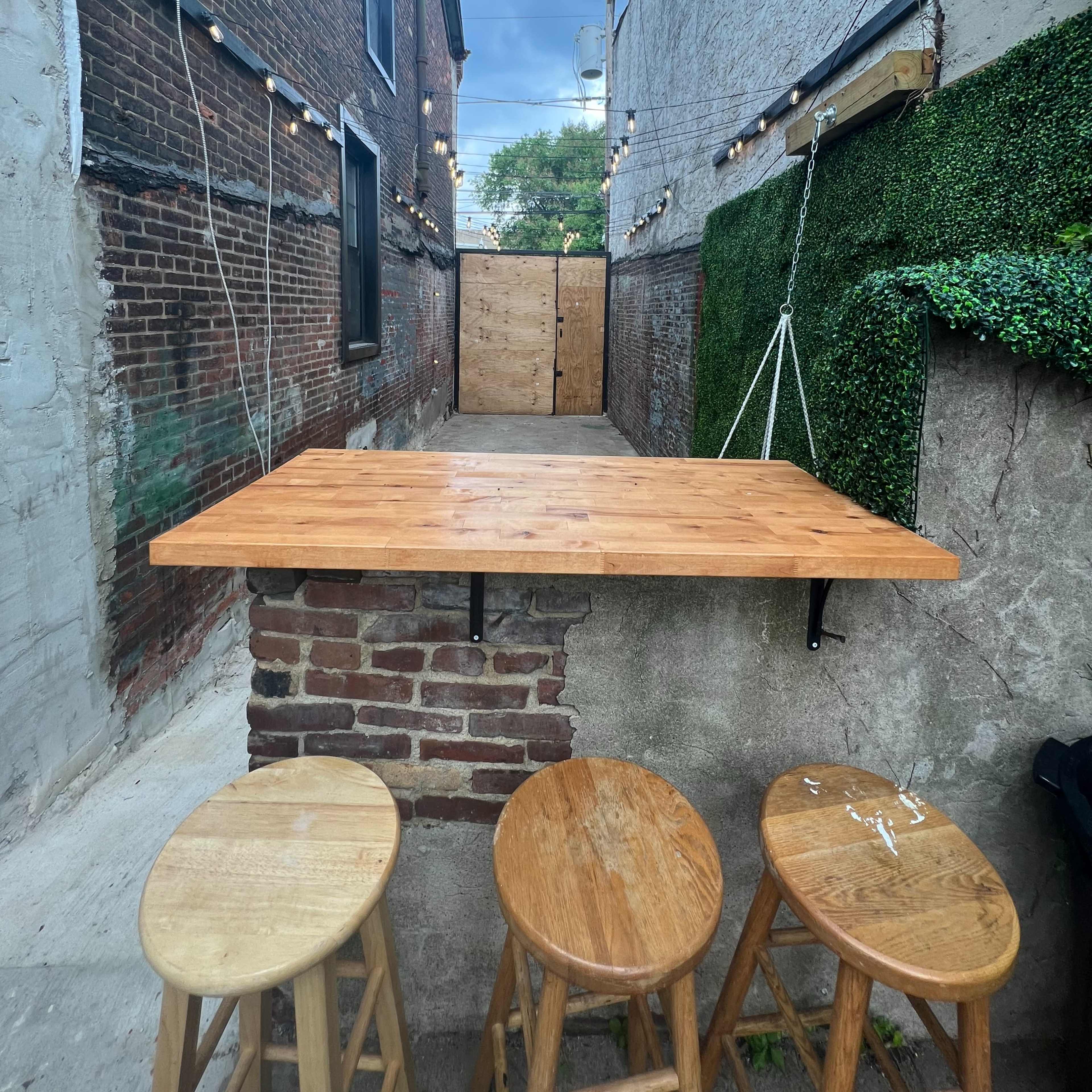 A wooden bar table with three stools is positioned in a narrow alley, flanked by brick and concrete walls, with greenery and string lights overhead.