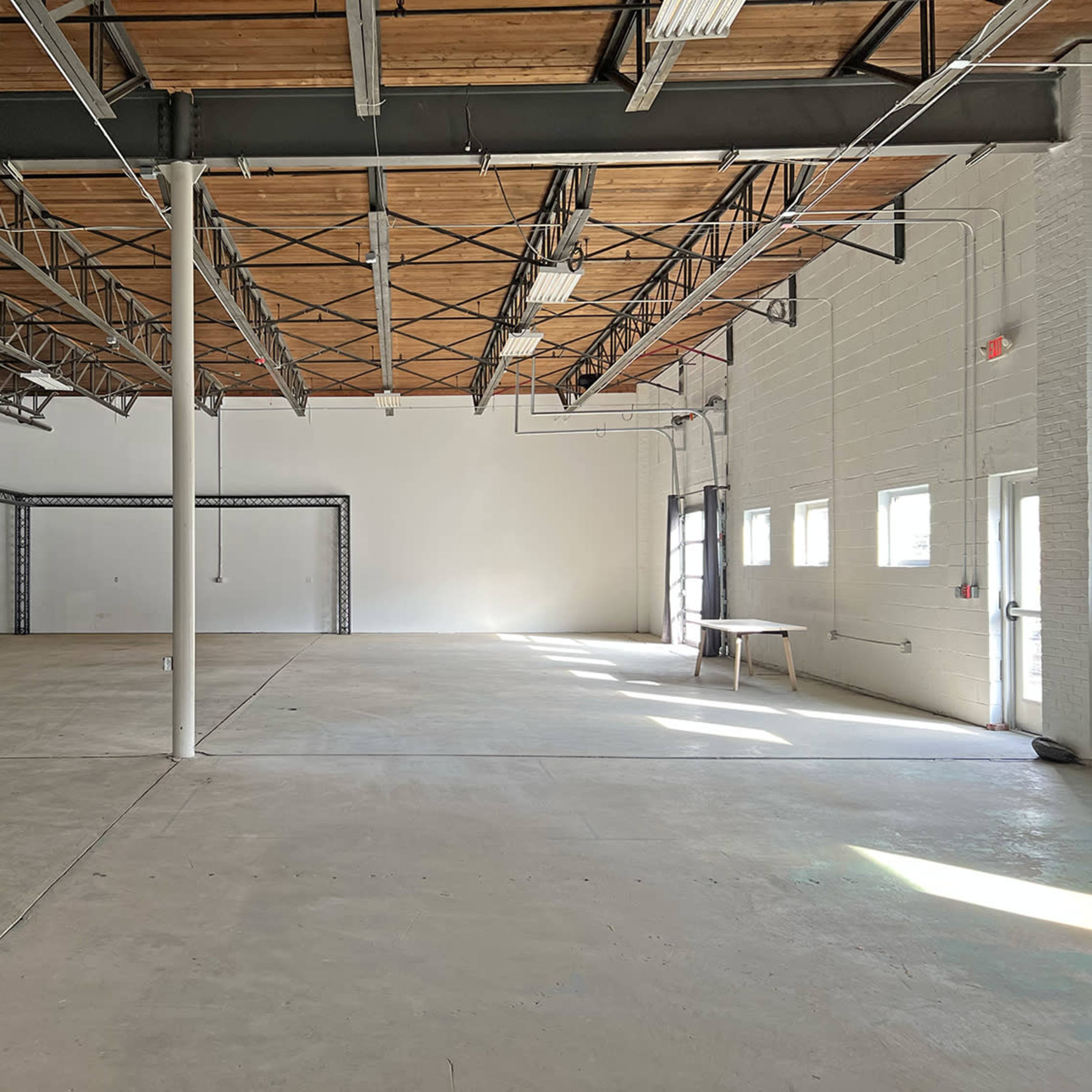 An empty industrial space with exposed beams, concrete flooring, and windows allowing natural light.