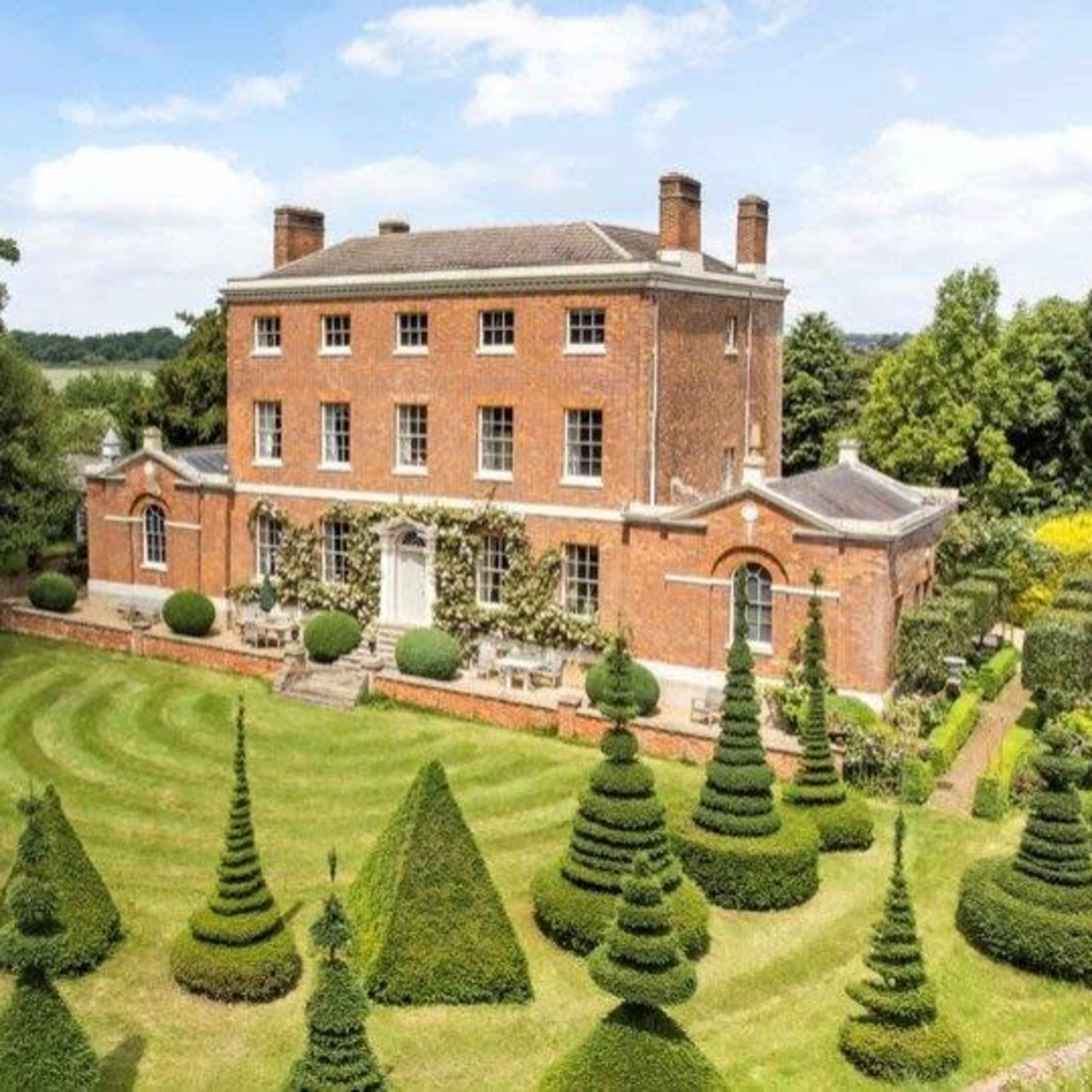 A large red brick mansion is surrounded by meticulously trimmed topiary in a landscaped garden.