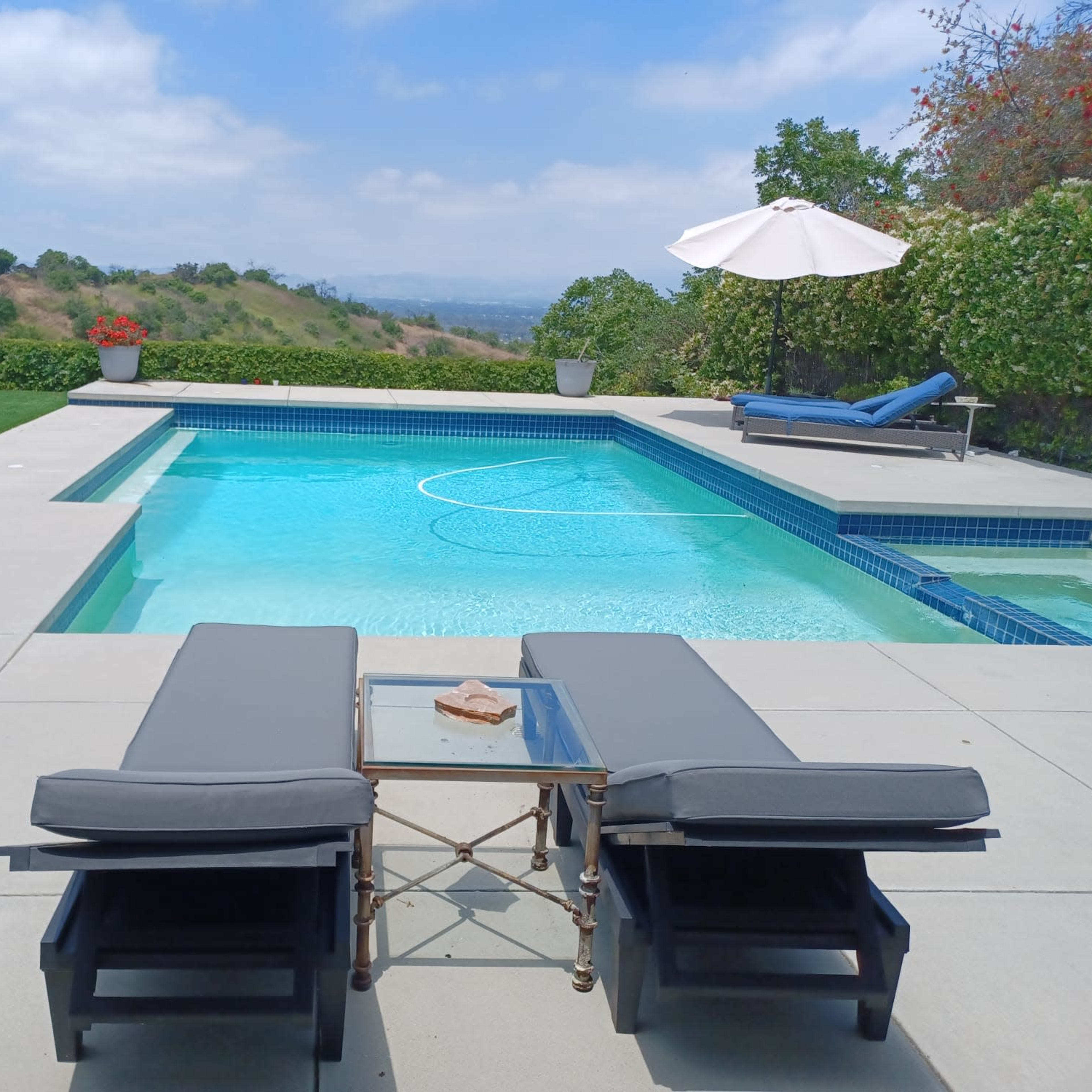 A swimming pool with chaise lounges and a side table is set against a backdrop of green hills under a partly cloudy sky.