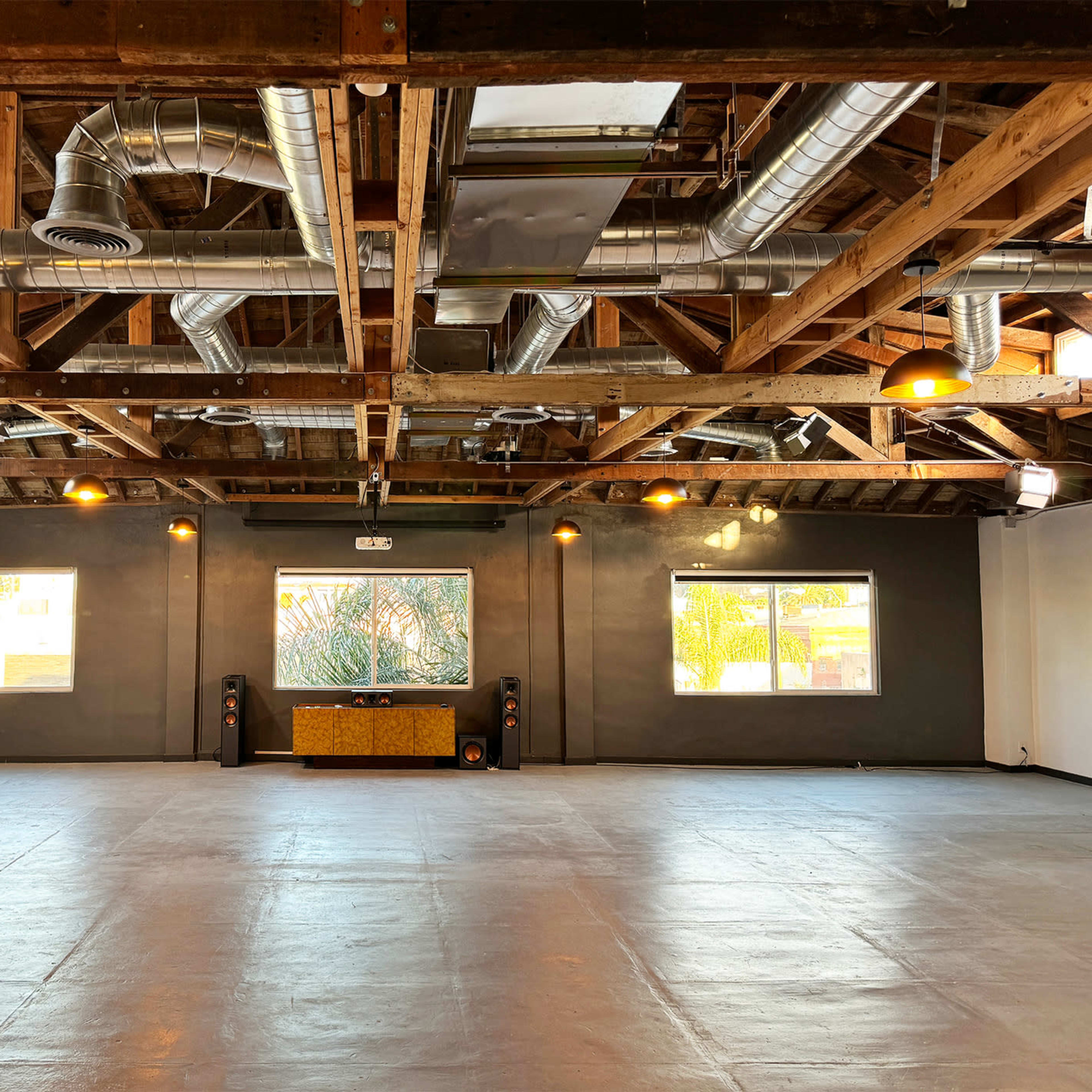 An empty studio space with exposed wooden beams, large windows, and minimal furniture, featuring a sound system against one wall.